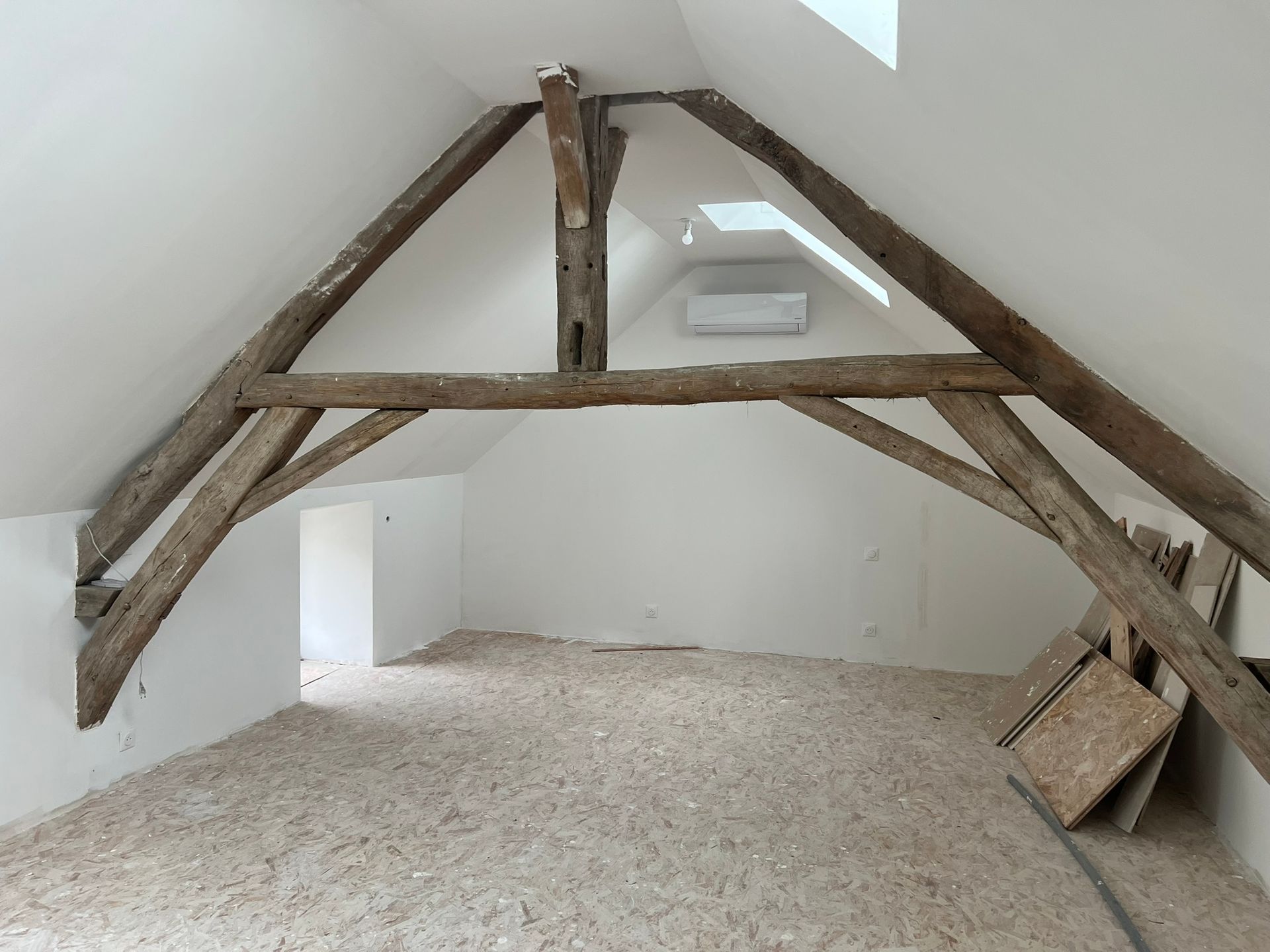 Chambre mansardée avec poutres apparentes, murs blancs et sol clair.