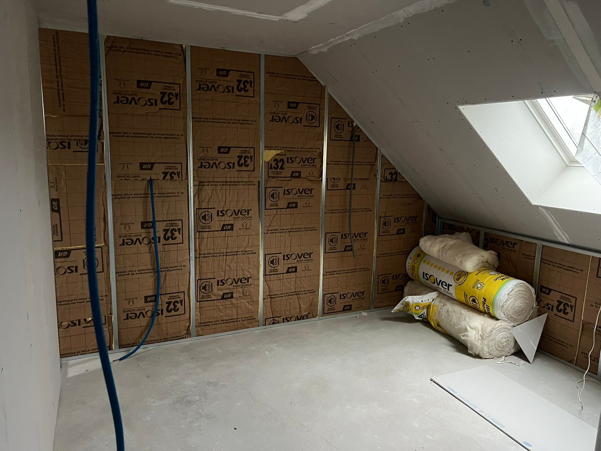 Pièce en construction, murs isolés avec un matériau brun. Quelques matériaux et une fenêtre sont présents.