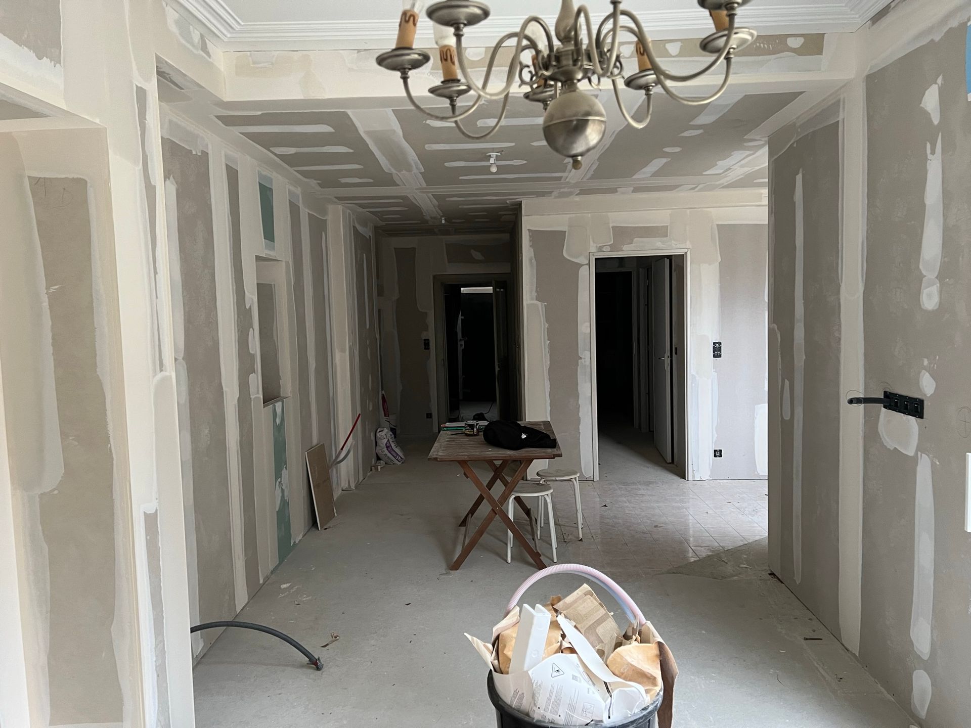Pièce en cours de rénovation ; murs et plafond recouverts de plaques de plâtre ; lustre suspendu ; débris dans un seau.