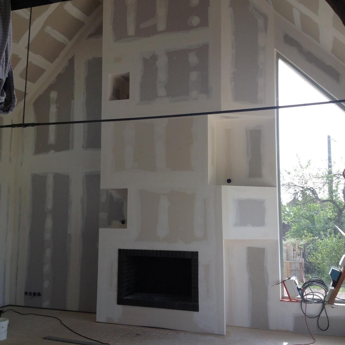 Intérieur en cours d'aménagement avec cheminée et cloisons sèches. Une fenêtre donne sur l'extérieur.