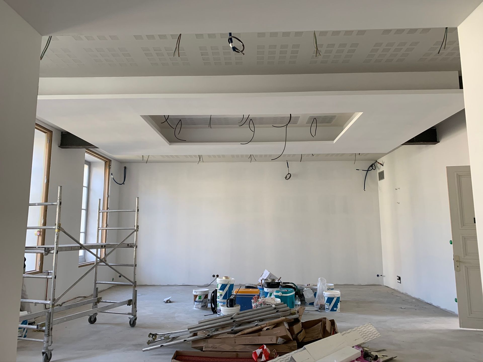 Vue intérieure d'une pièce en construction avec un plafond en retrait, des échafaudages et des câbles électriques.