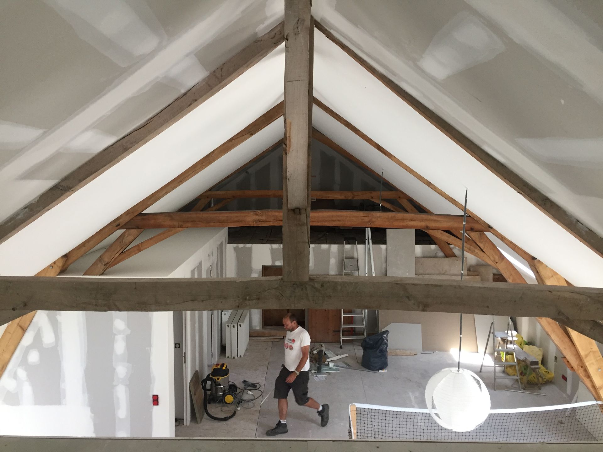 Vue intérieure d'un bâtiment avec poutres en bois apparentes, cloisons sèches et une personne marchant au niveau inférieur.