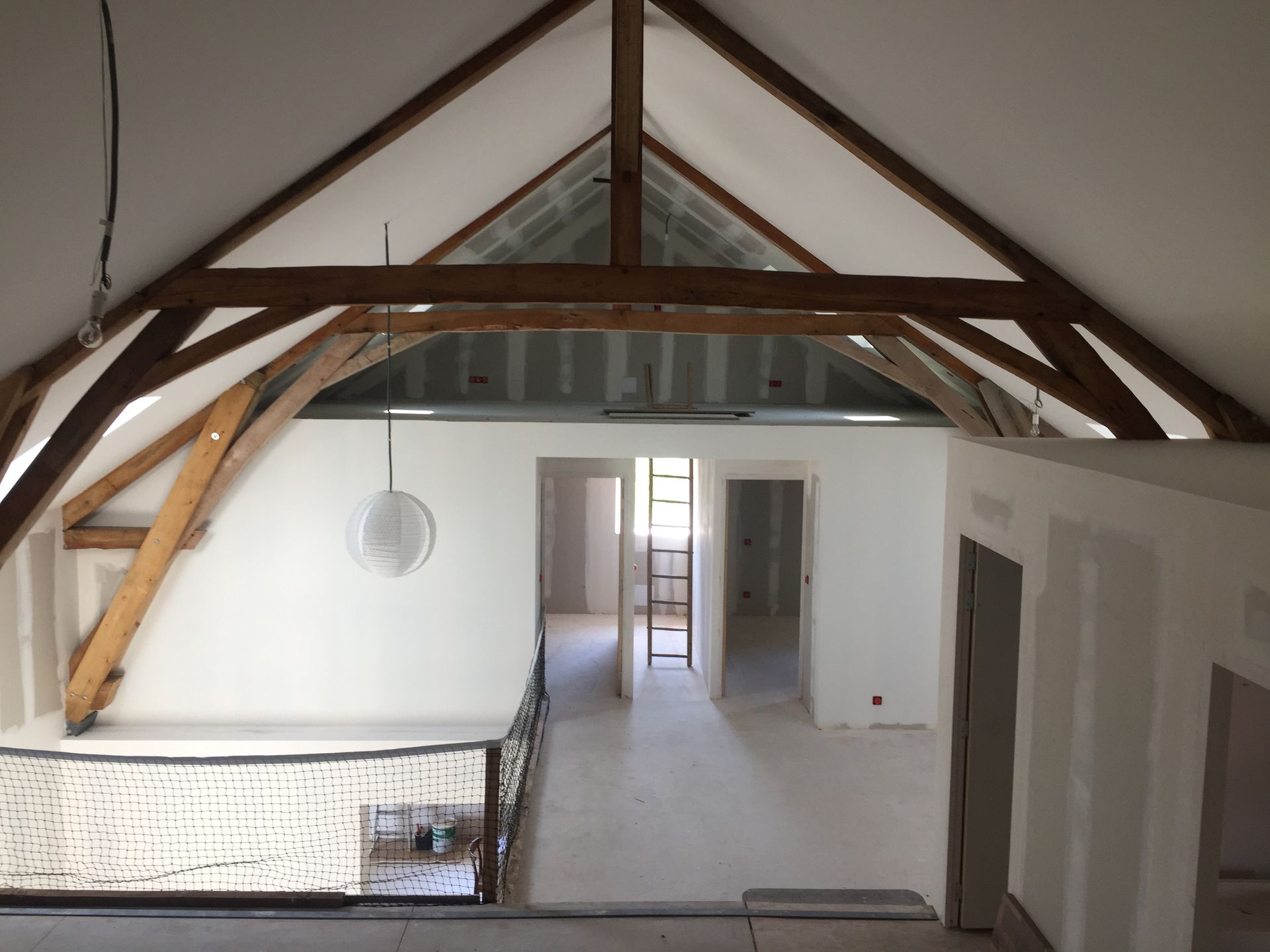 Intérieur d'une maison en construction avec poutres en bois apparentes, portes ouvertes et balustrade au deuxième étage.