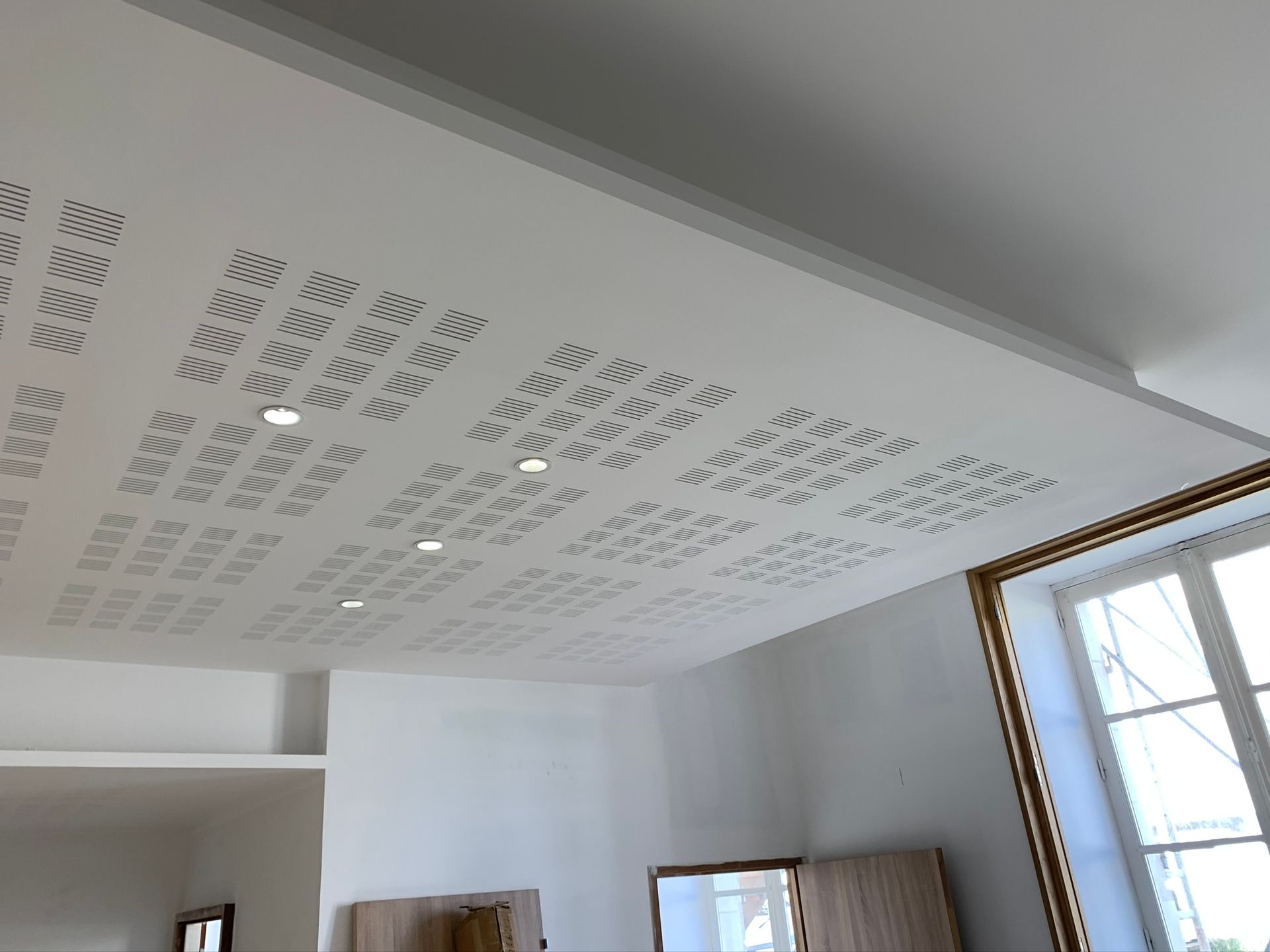 Panneau de plafond blanc perforé avec éclairage encastré, adjacent à une fenêtre.