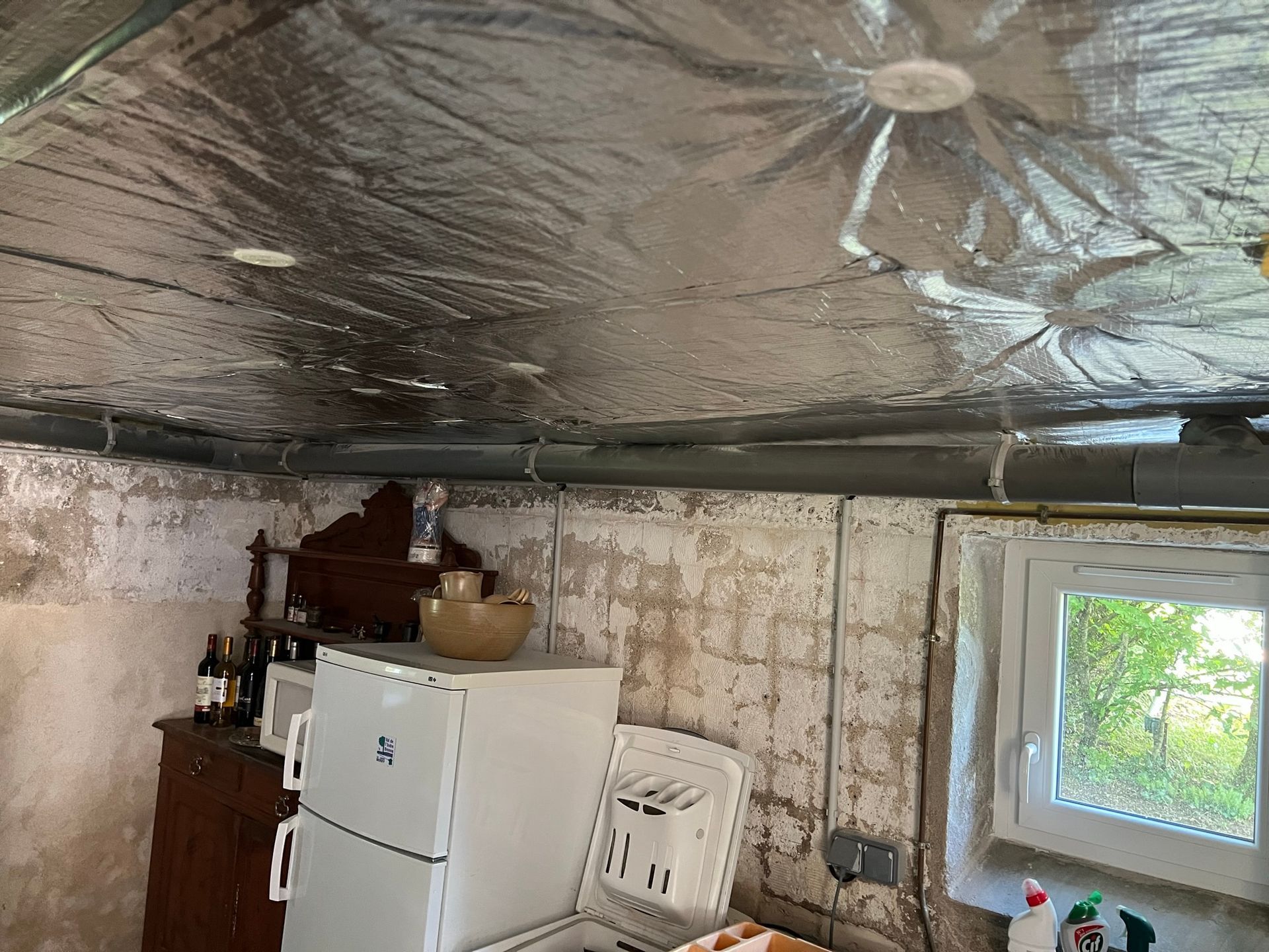 Cuisine au sous-sol avec plafond recouvert de papier aluminium, réfrigérateur blanc, petite fenêtre et murs en béton.