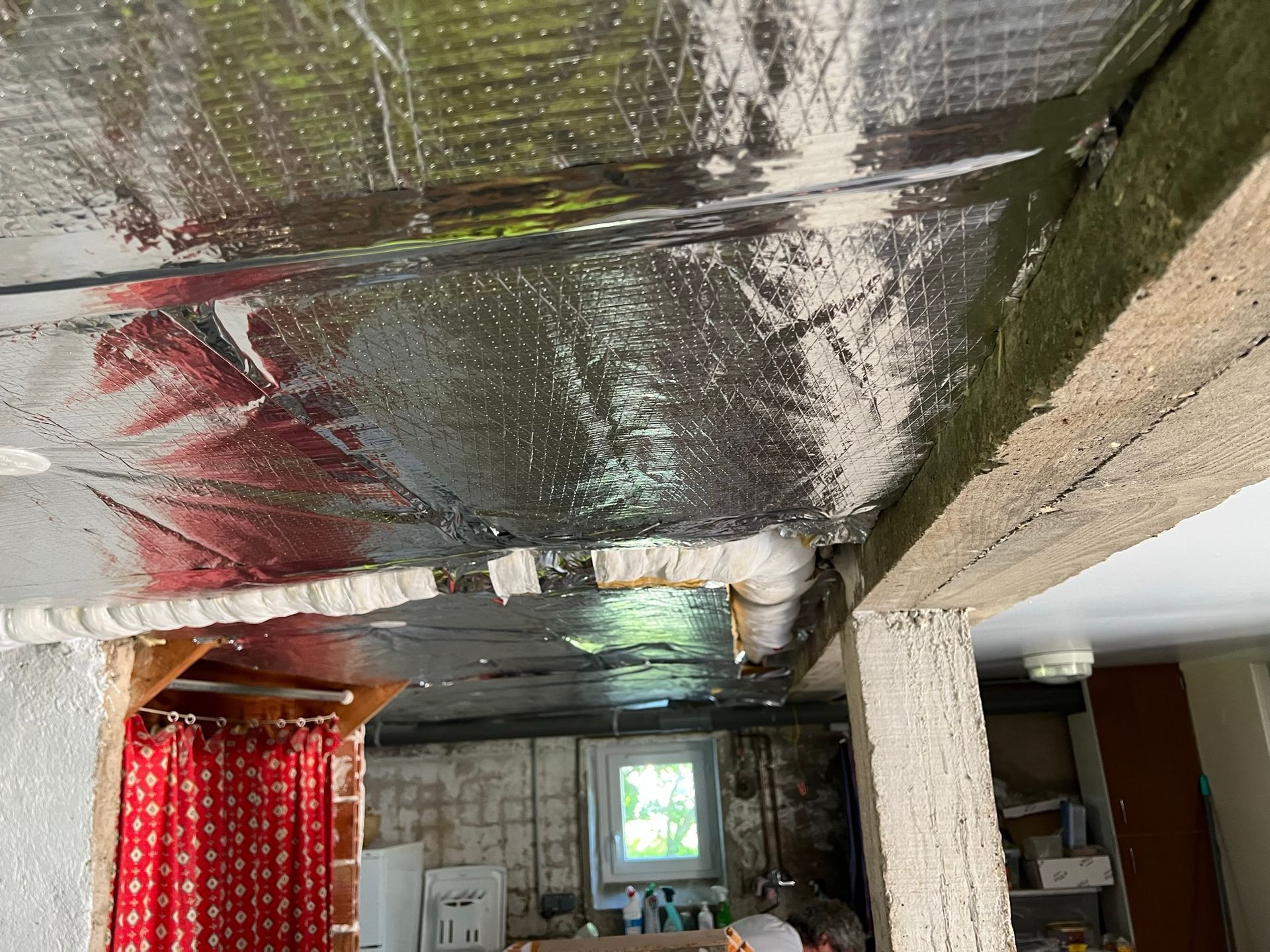 Plafond isolé par une feuille d'aluminium, poutres en béton et aperçu d'une pièce avec une fenêtre et un rideau rouge.