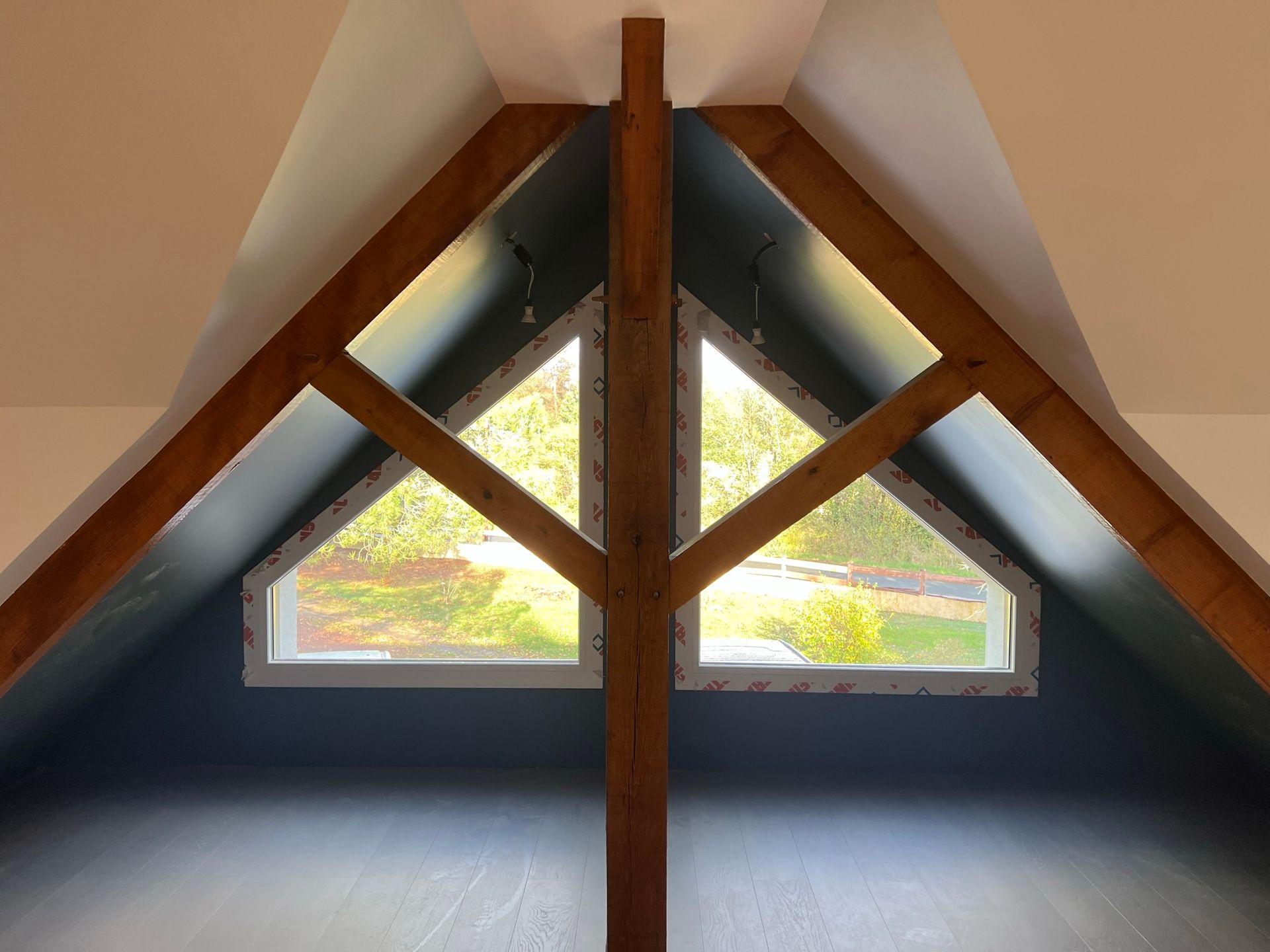 Vue intérieure d'une fenêtre triangulaire aux murs bleu foncé, poutres en bois et aperçu de verdure à l'extérieur.