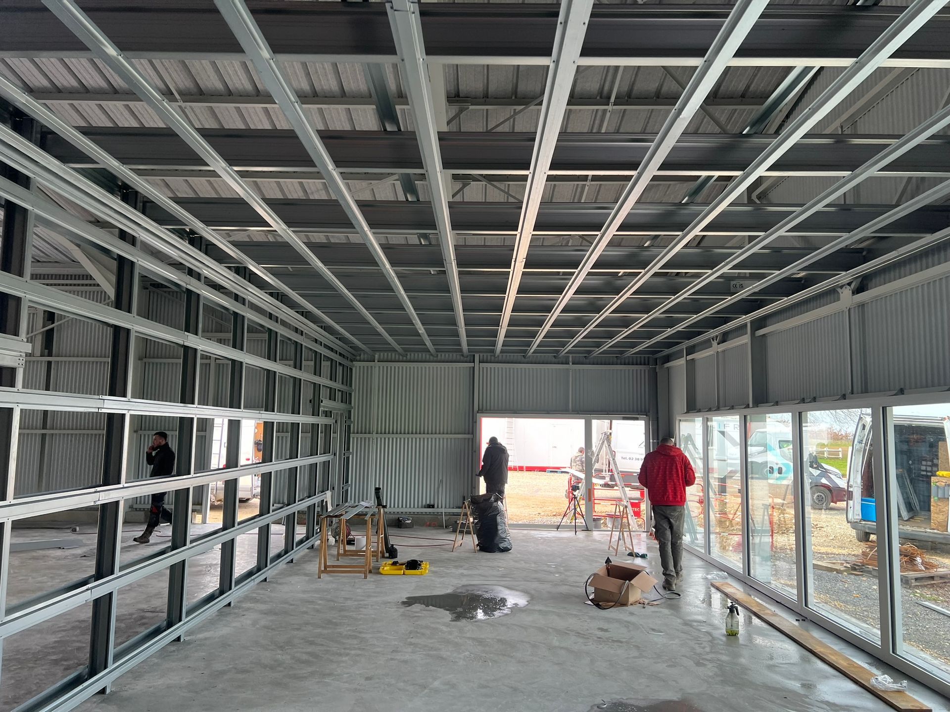 Intérieur d'un bâtiment en construction. Ossature métallique pour les murs et le plafond. Passage de personnes. Sol en béton.