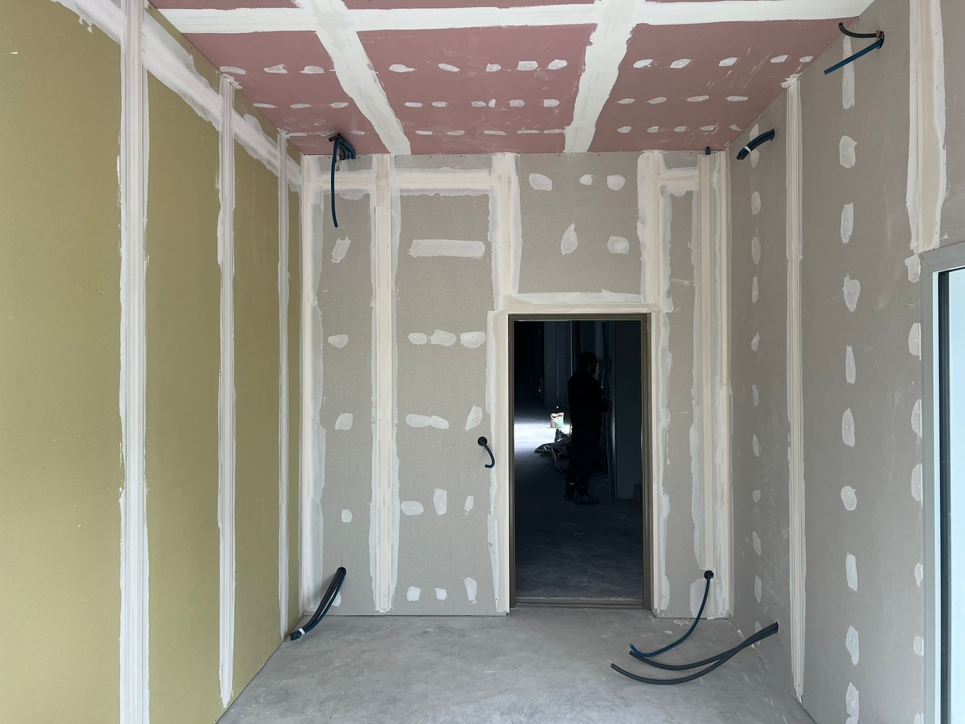 Pièce intérieure en travaux, avec cloisons sèches apparentes, plafond non fini et porte. Câbles électriques visibles.