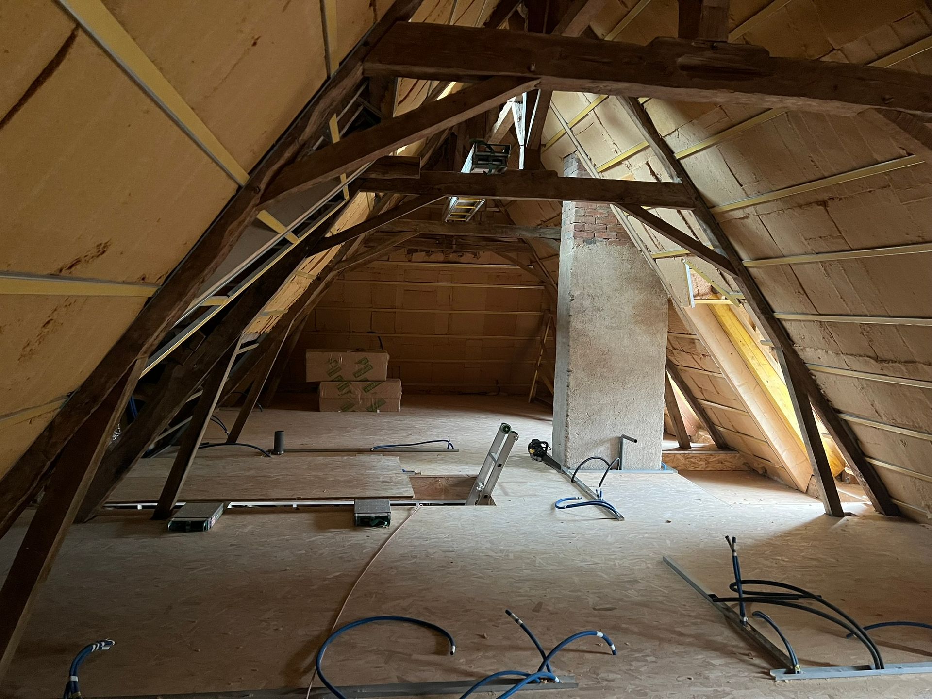 Intérieur d'un grenier non aménagé avec poutres en bois, murs isolés et câbles électriques apparents.