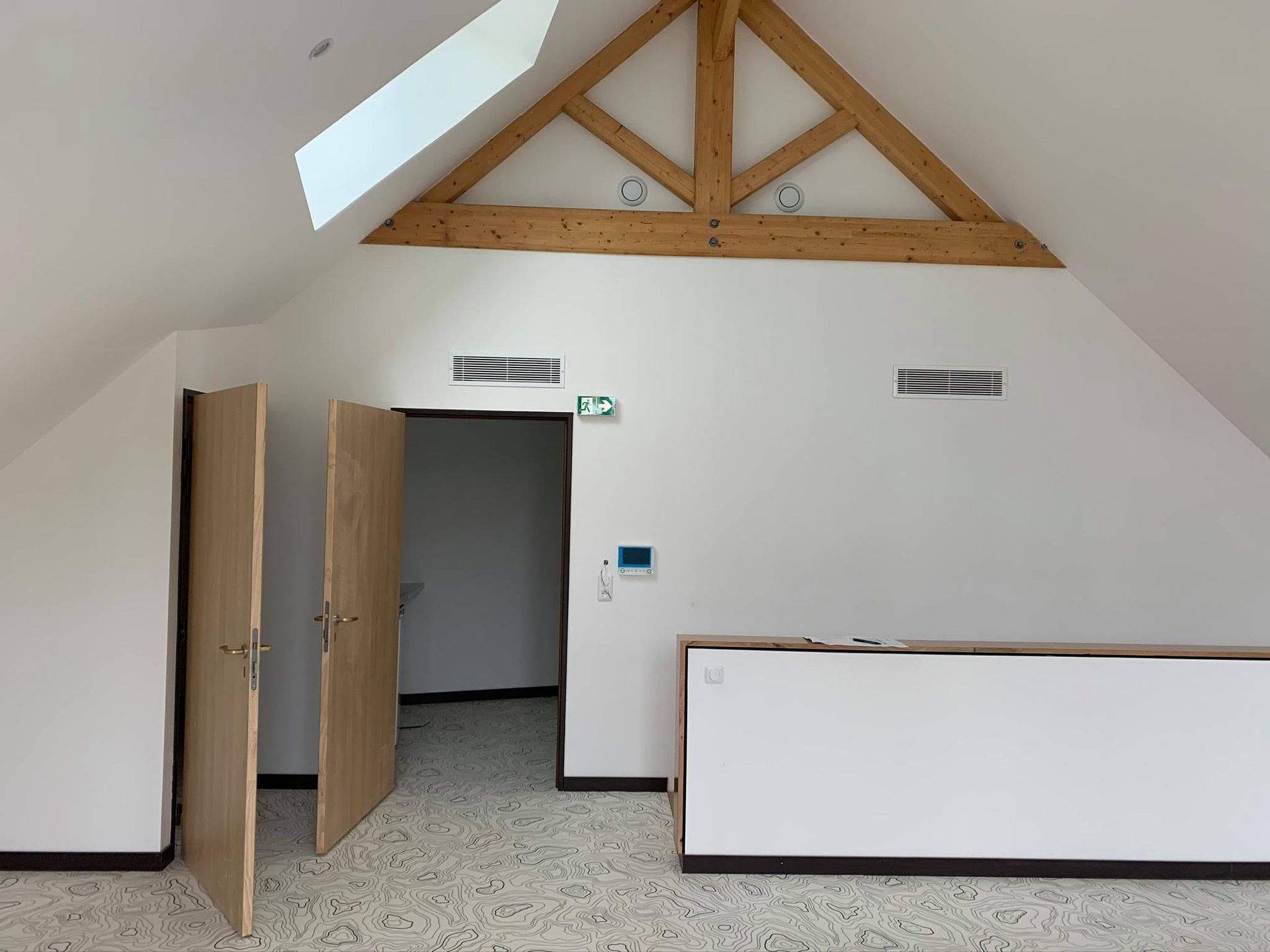 Intérieur d'une pièce mansardée avec poutres en bois, deux portes, une entrée et un mur blanc.
