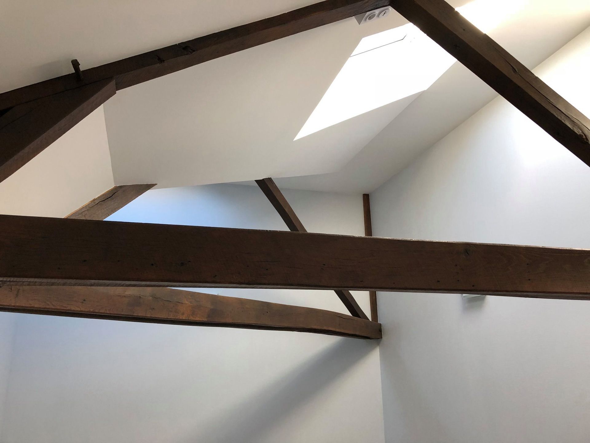Intérieur d'une pièce avec poutres apparentes, murs blancs et puits de lumière.