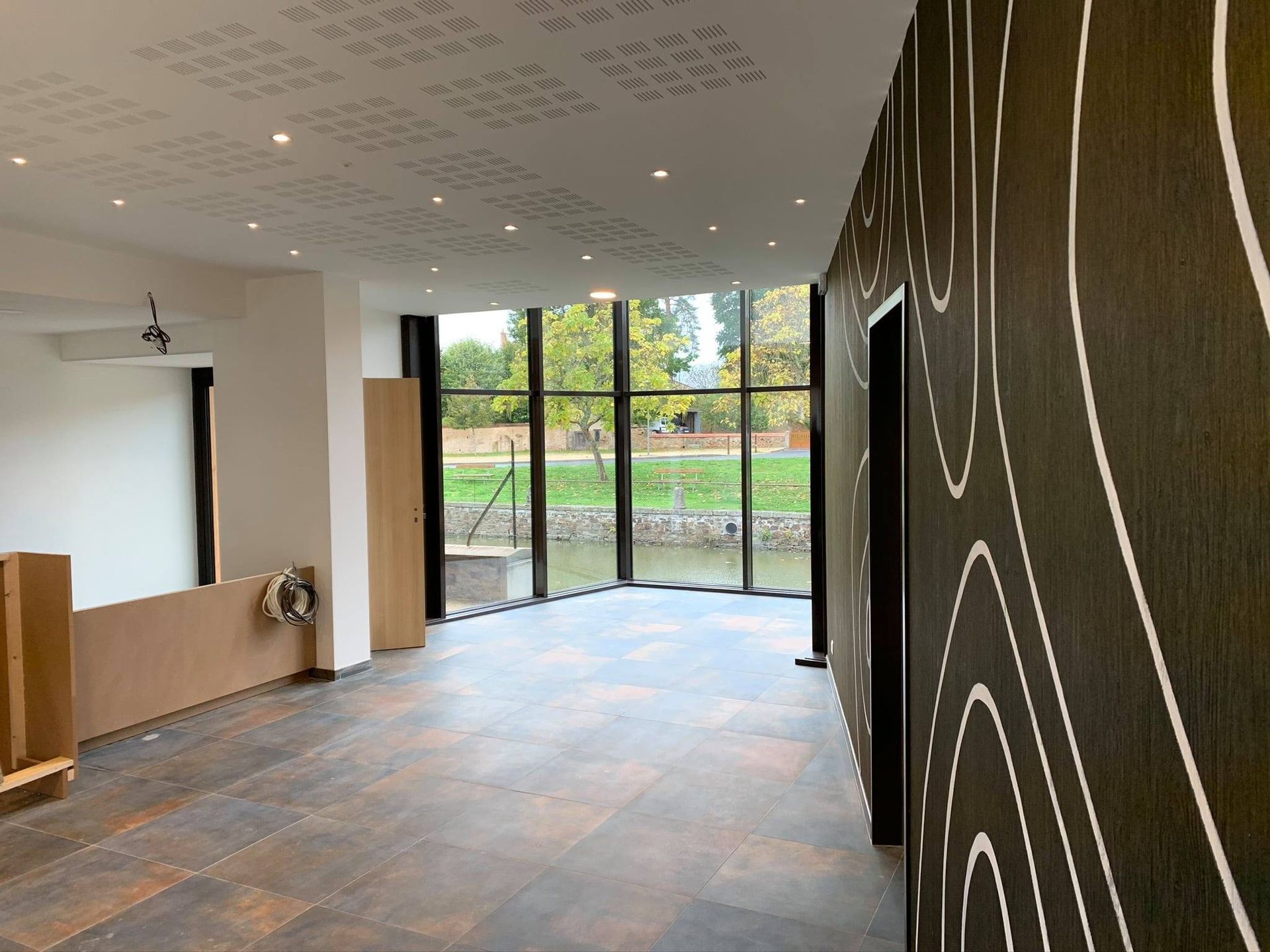 Pièce vide avec un mur à motifs sombres, une grande fenêtre donnant sur un cours d'eau et un éclairage au plafond.