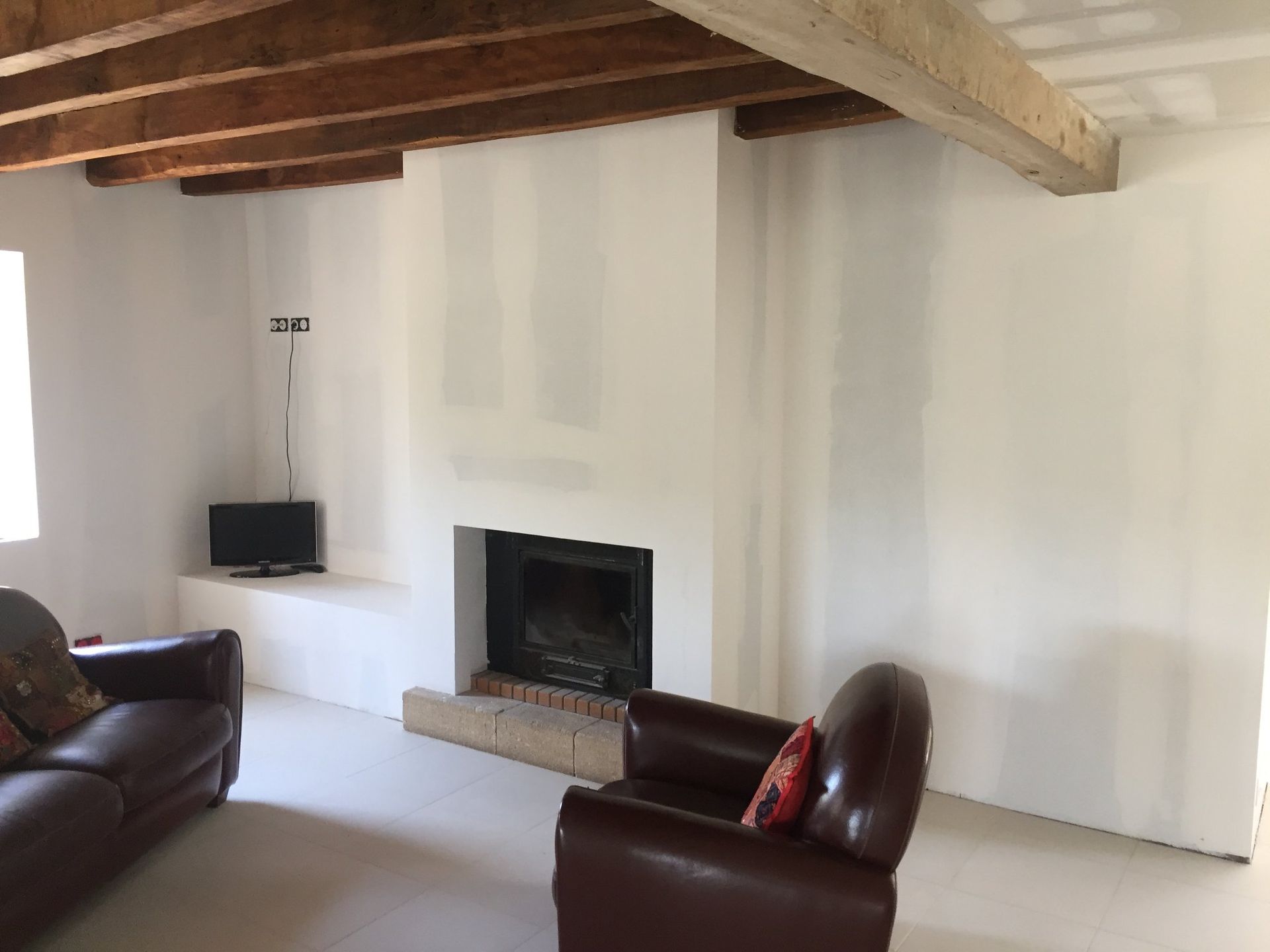 Salon avec cheminée, murs blancs, poutres apparentes et deux fauteuils en cuir.
