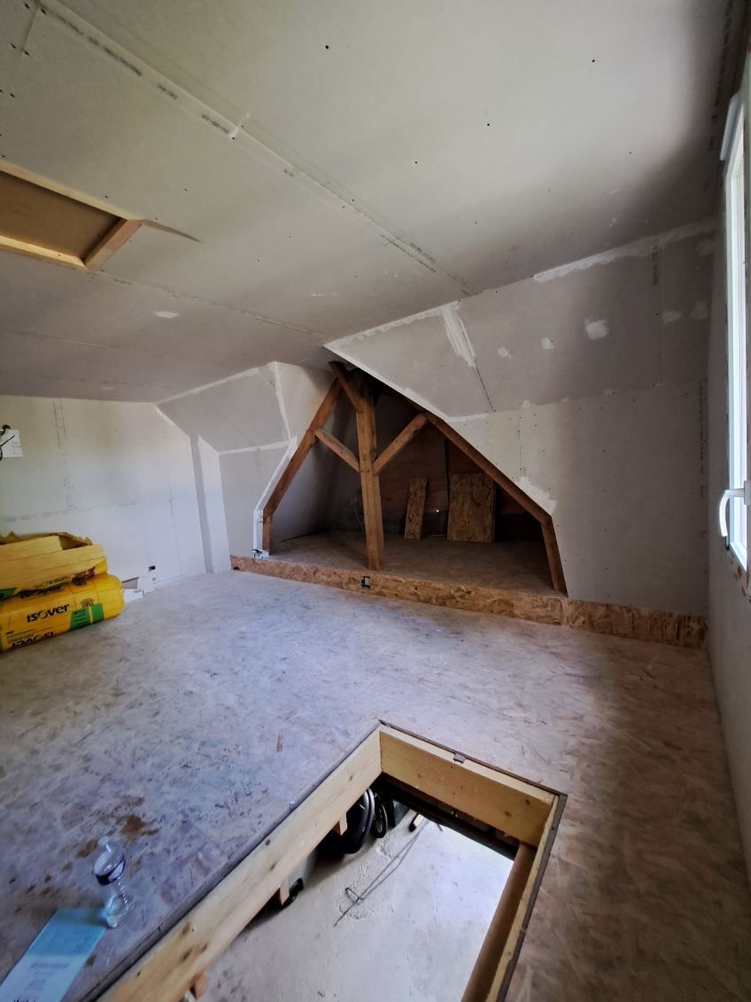 Vue intérieure d'un grenier non aménagé. Poutres et chevrons apparents, plaques de plâtre aux murs et au plafond.