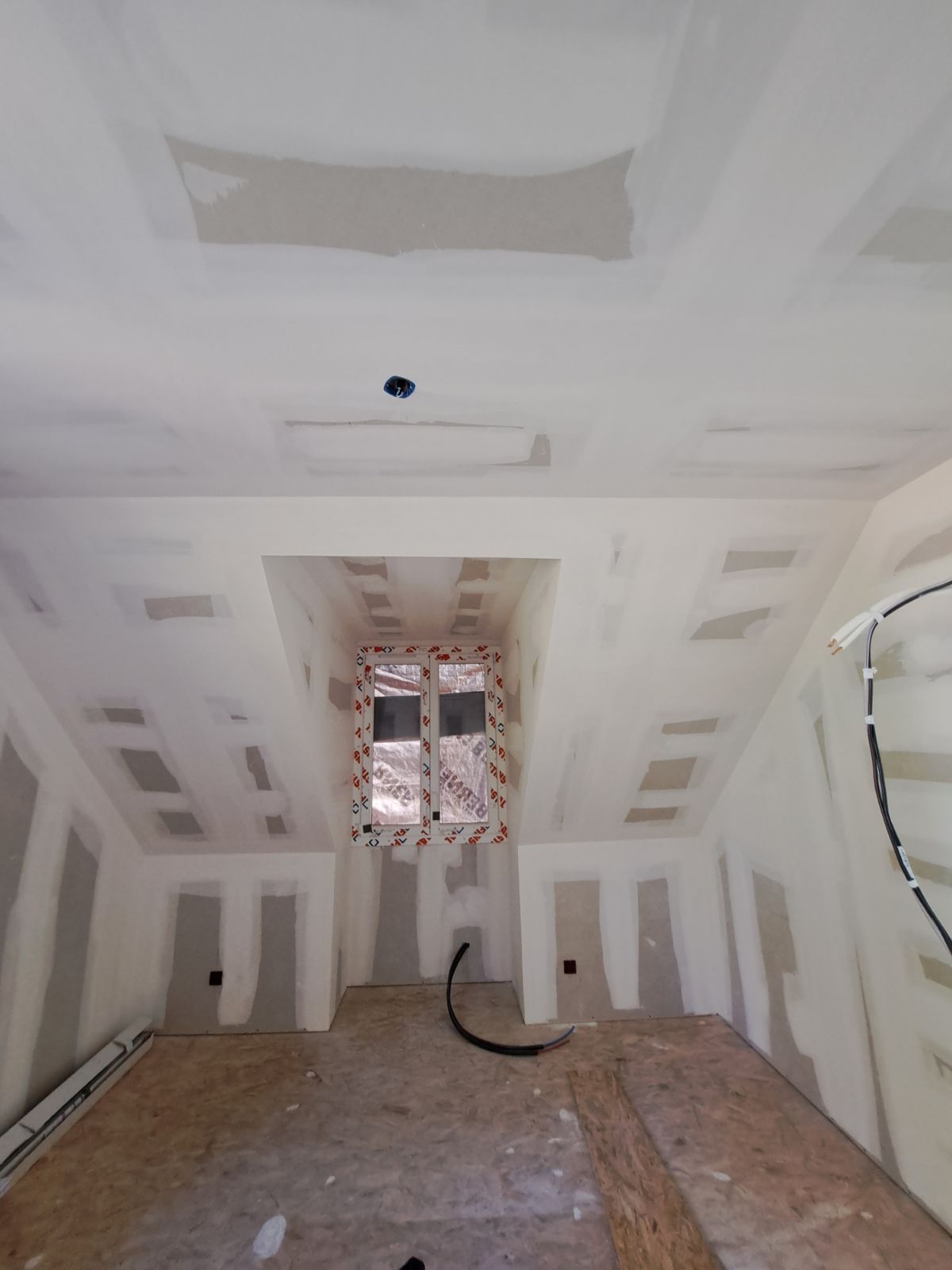 Pièce intérieure en cours d'aménagement, avec des plaques de plâtre installées aux murs et au plafond, fenêtre au centre.