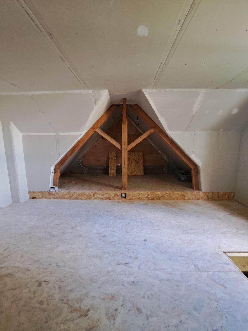 Vue intérieure d'un grenier non aménagé avec poutres apparentes et cloisons sèches. Sol et murs de couleur claire.