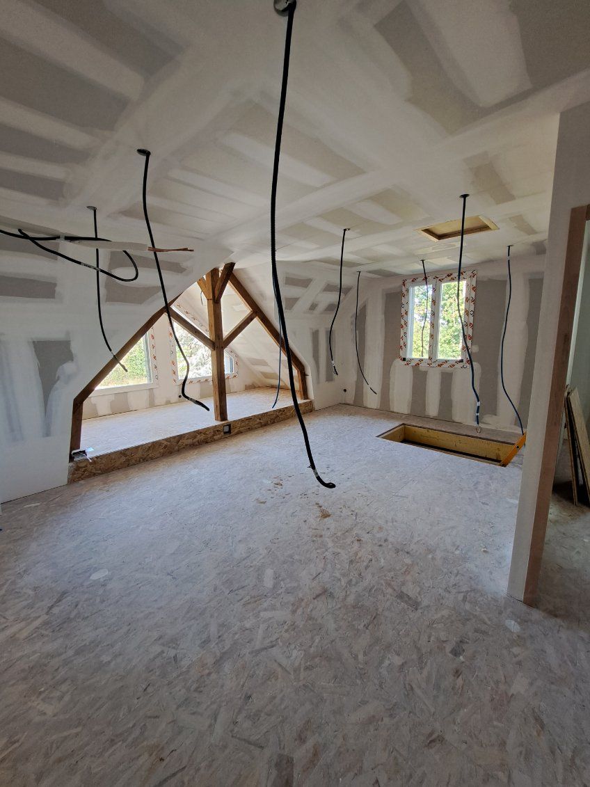 Pièce en travaux avec poutres apparentes, câblage et isolation au sol. Tons clairs et neutres.