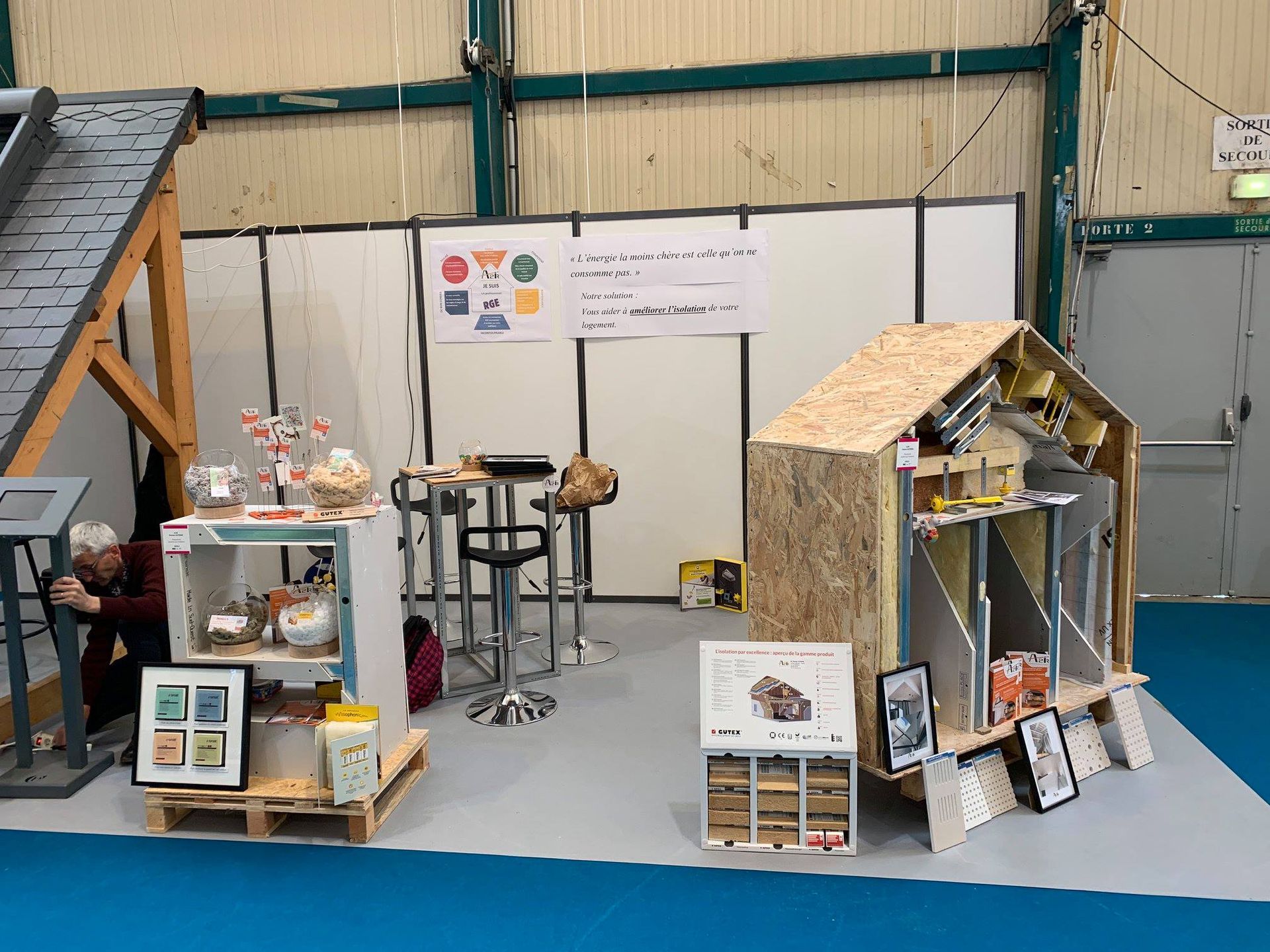 Stand d'exposition : maison miniature, présentation de produits, sol gris et fond blanc.