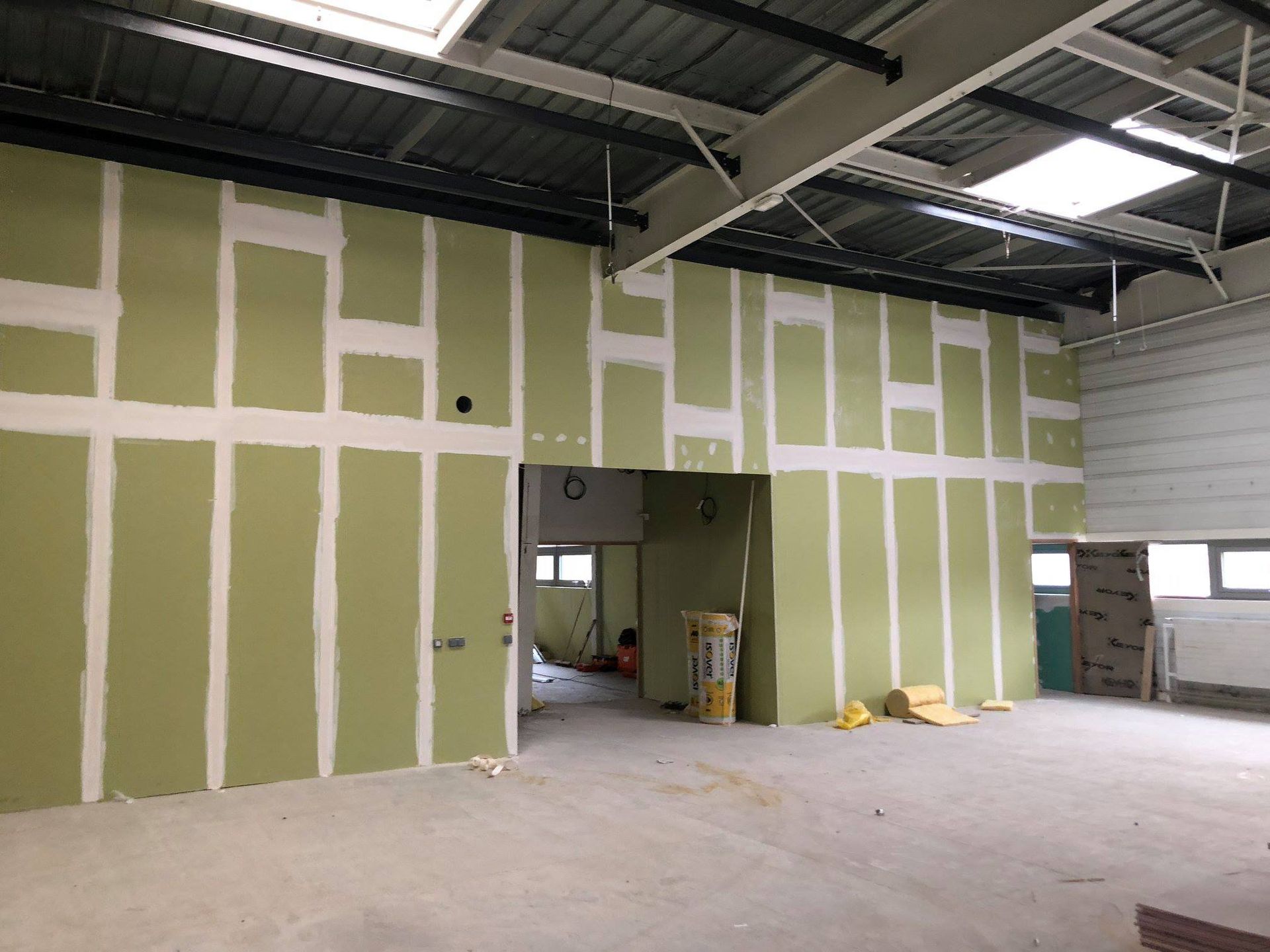 Vue intérieure d'un bâtiment en construction. Murs en plaques de plâtre vertes, poutres métalliques et sols en béton.