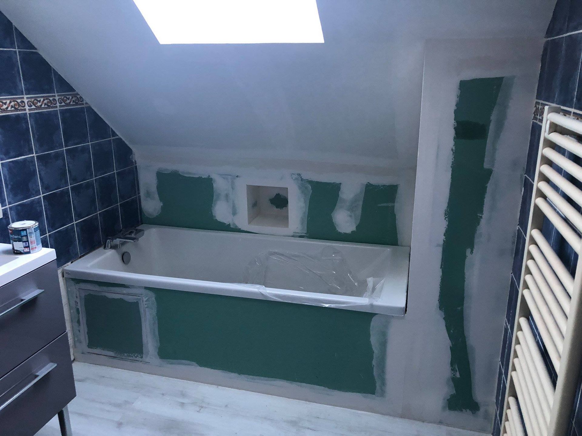 Salle de bains avec baignoire sous un plafond mansardé ; cloisons sèches turquoise, carrelage bleu et baignoire blanche.