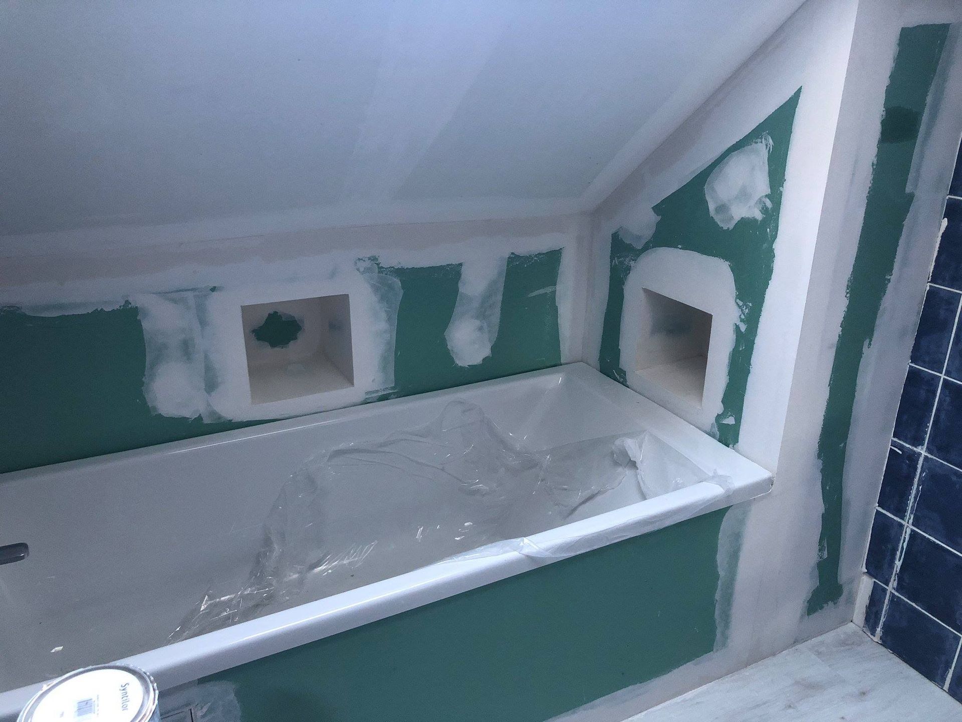 Une salle de bains partiellement aménagée avec baignoire et rangements intégrés, cloisons sèches vertes et  joints blancs.