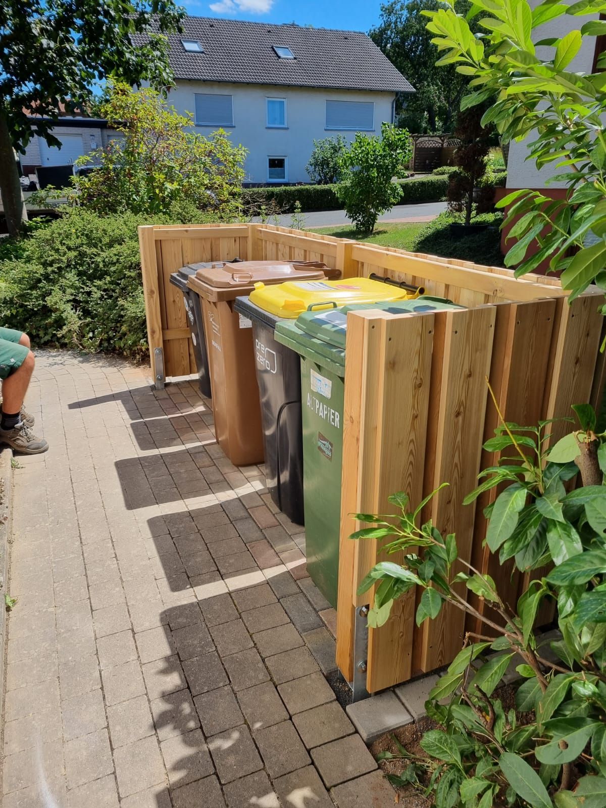 Neben einem Holzzaun steht eine Reihe Mülltonnen.