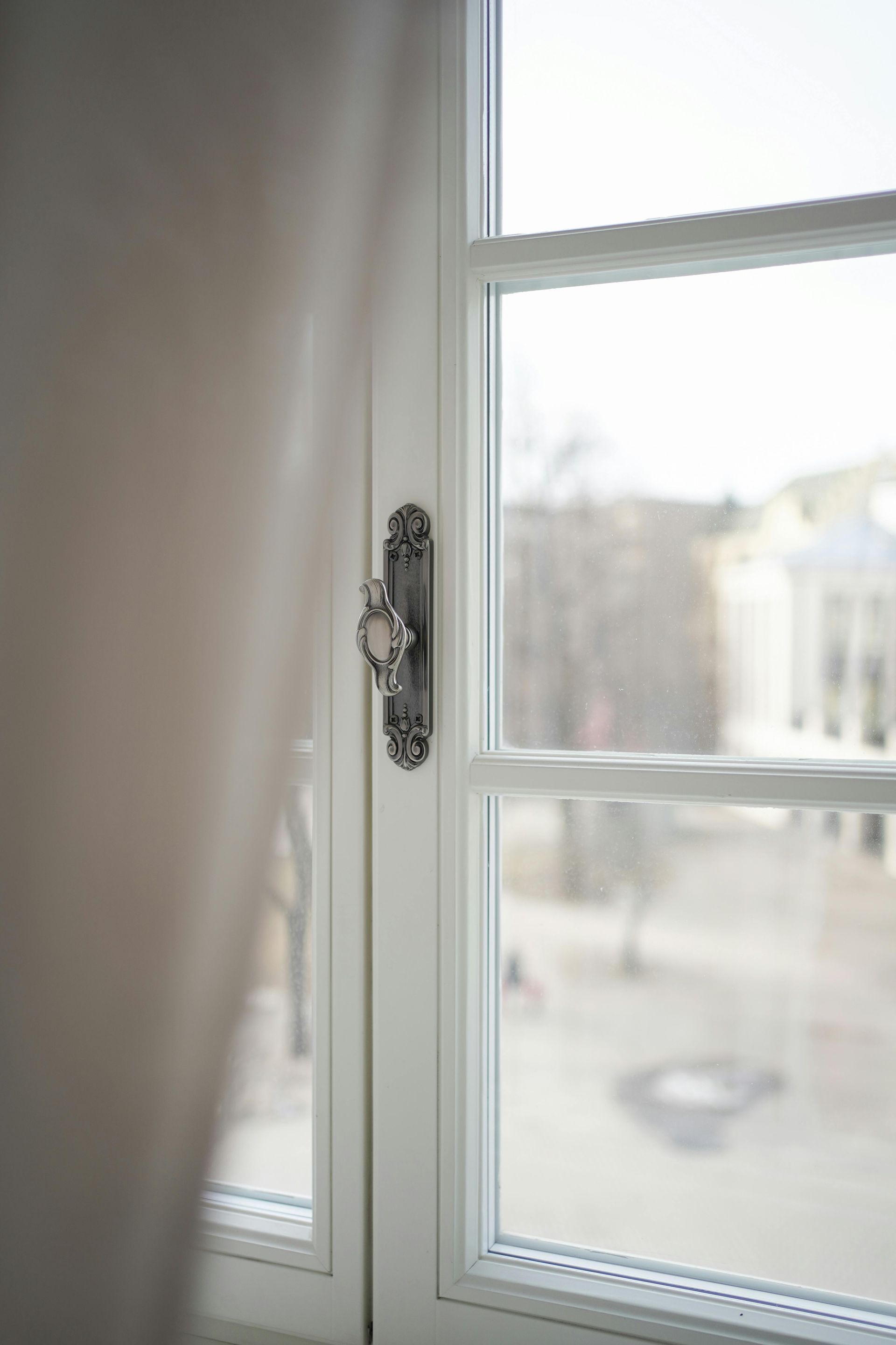Ein durchsichtiger Vorhang neben einem weißen Fenster mit einem verzierten Metallverschluss, sanftes Tageslicht draußen