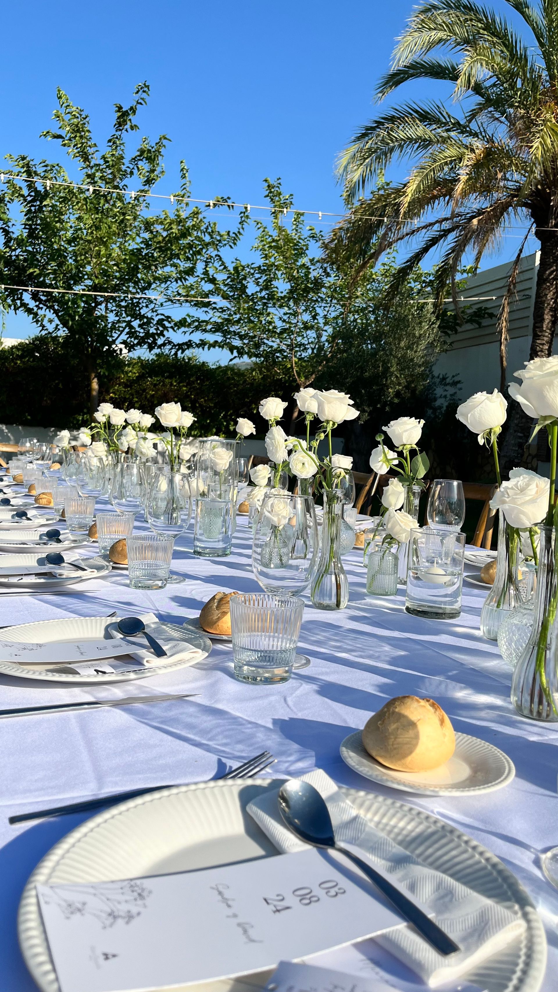 Una mesa larga con platos, utensilios, flores y una palmera al fondo.