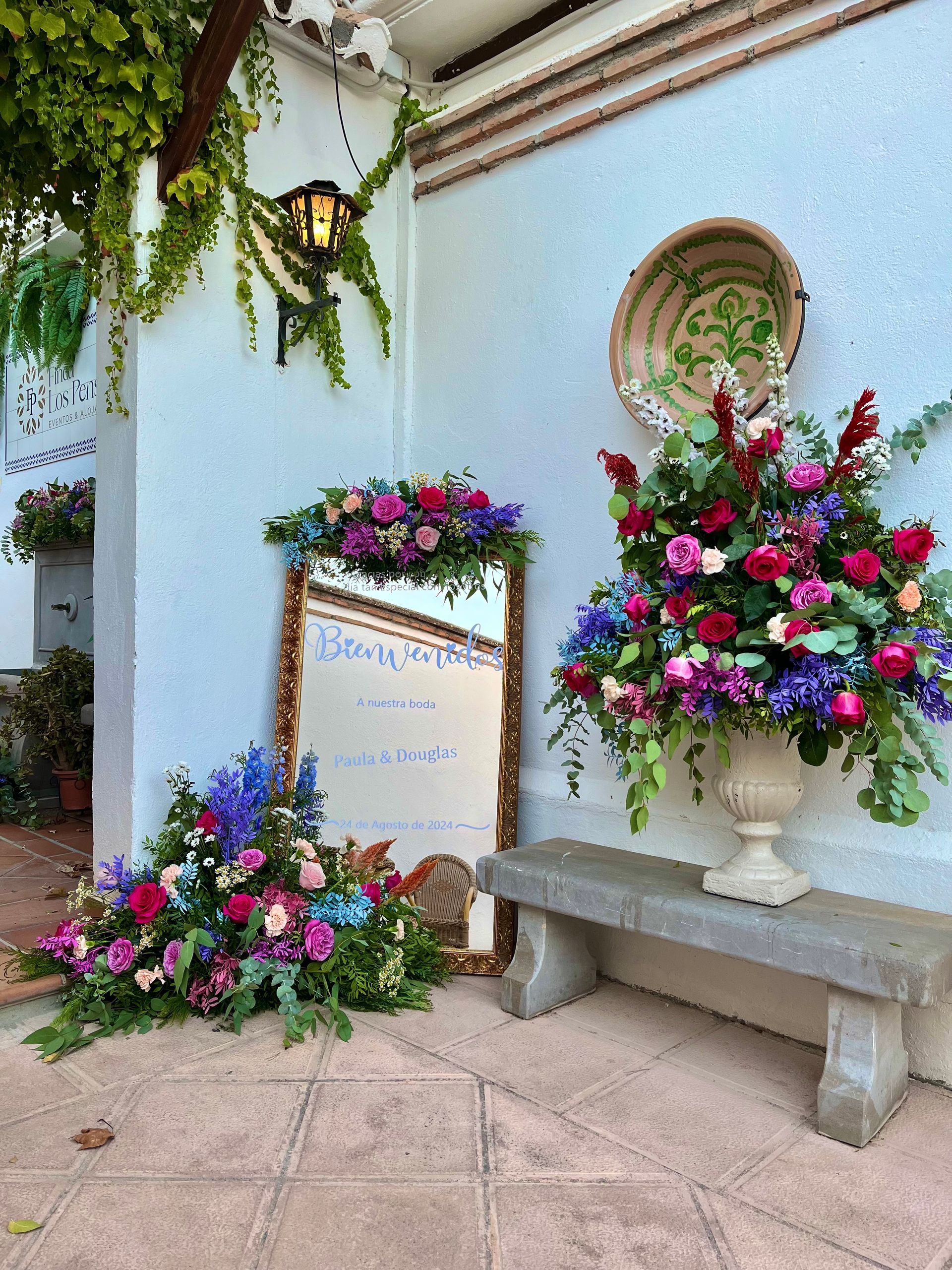 Un jarrón de flores está sobre un banco al lado de un espejo.