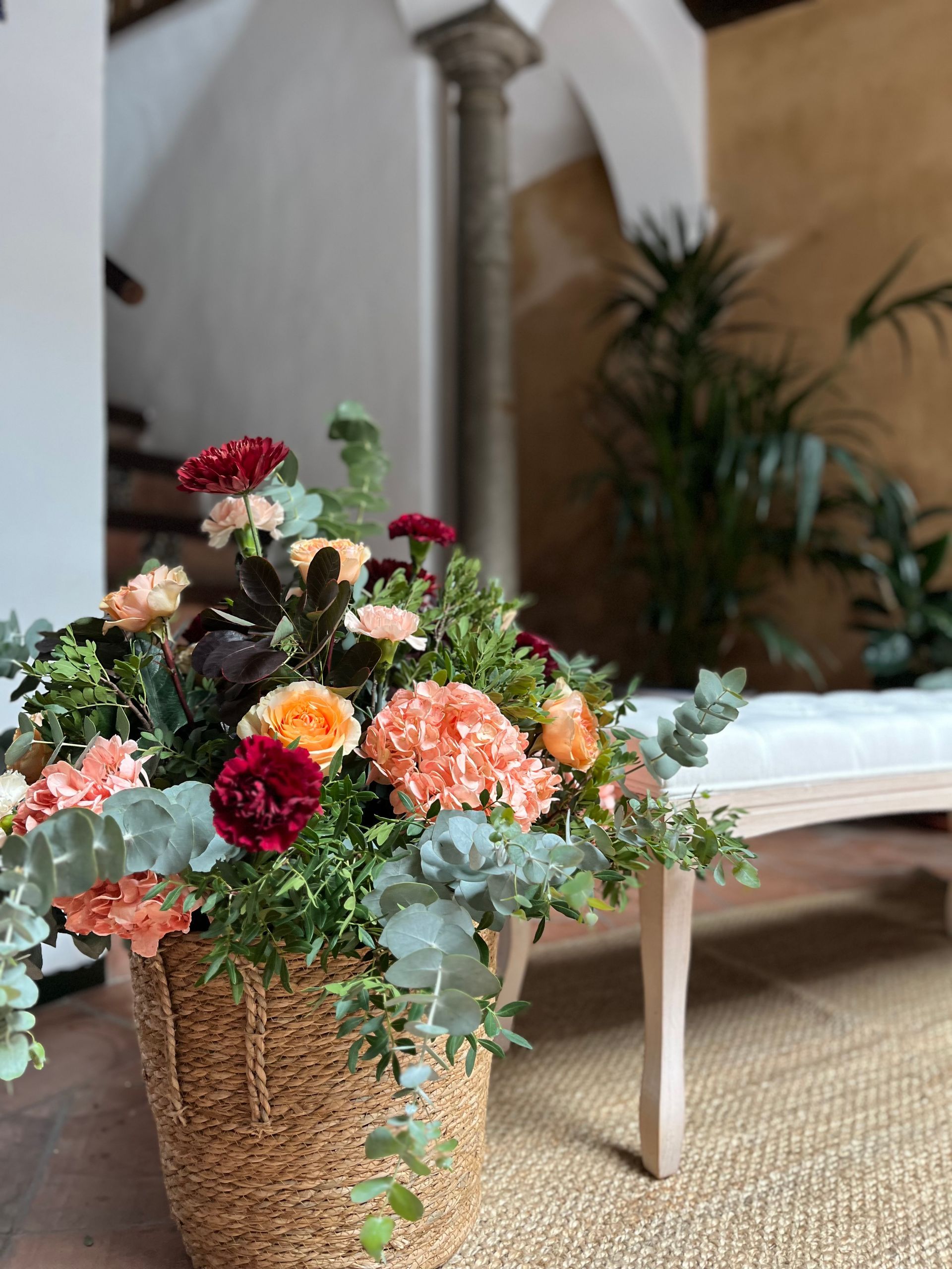 Una cesta llena de flores está en el suelo al lado de un banco.