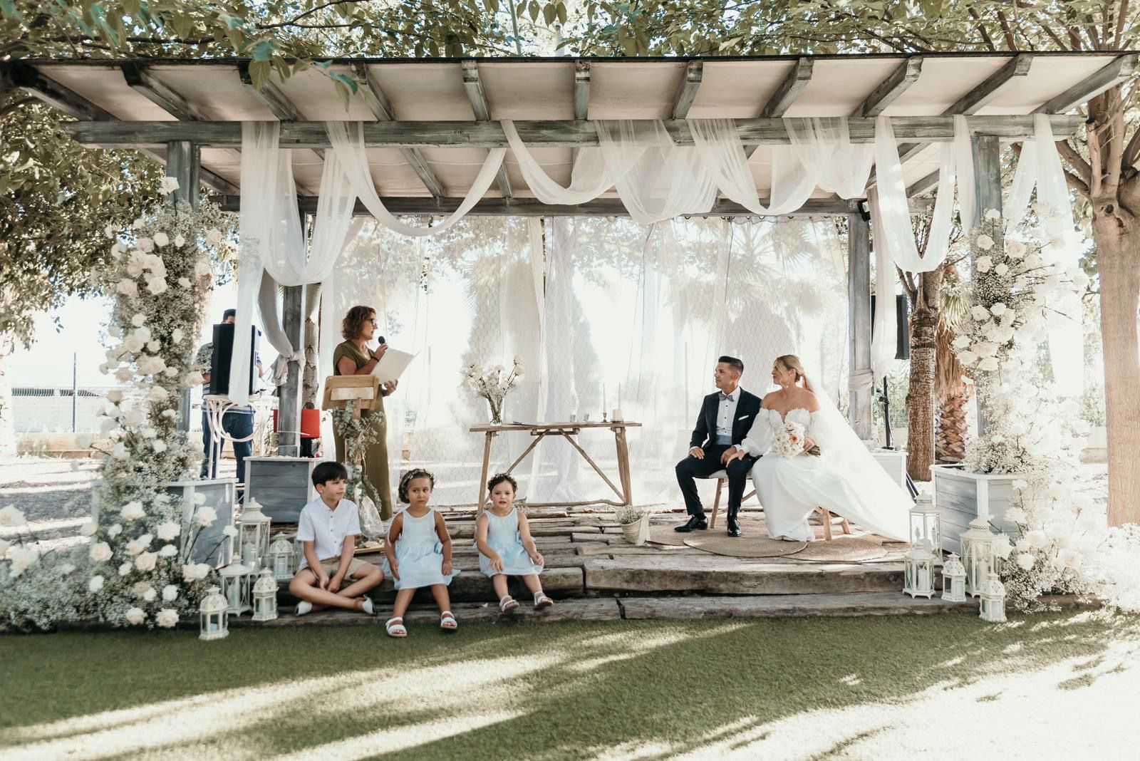 Una novia y un novio están sentados bajo una pérgola con sus hijos.