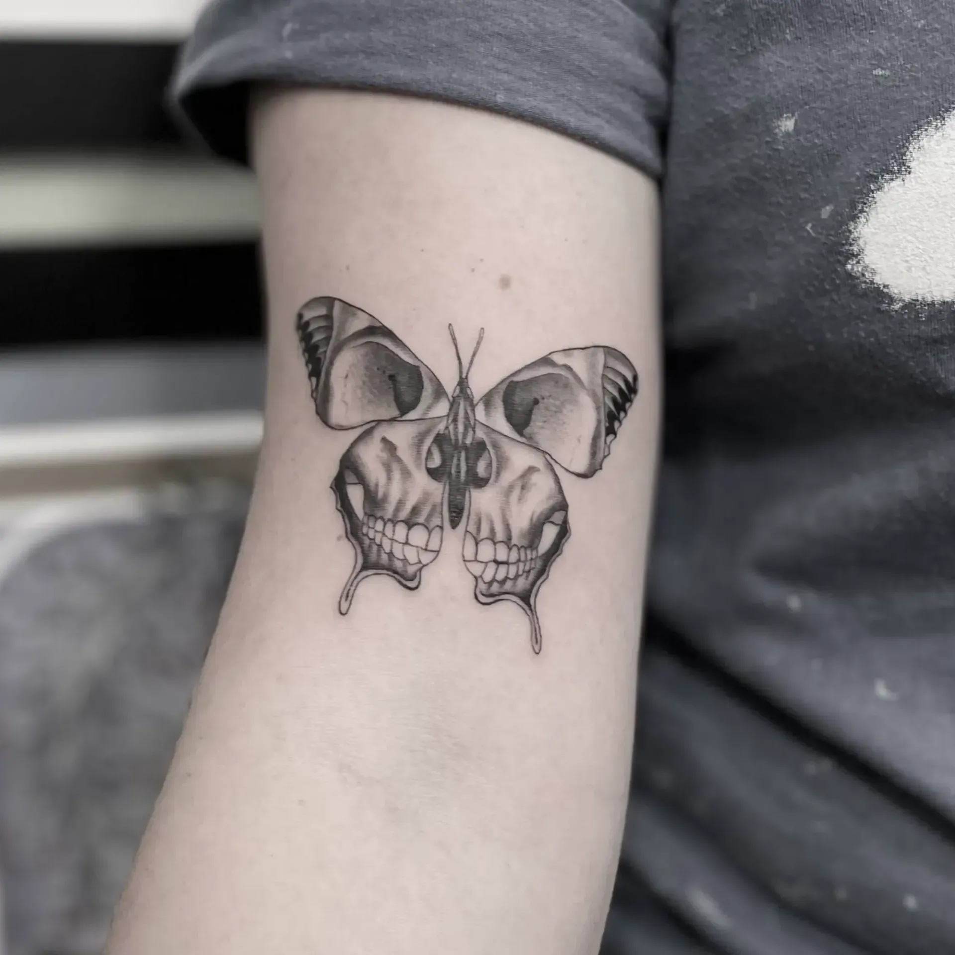 Un tatuaje en blanco y negro en un brazo que representa una mariposa cuyas alas tienen forma de cráneo humano.