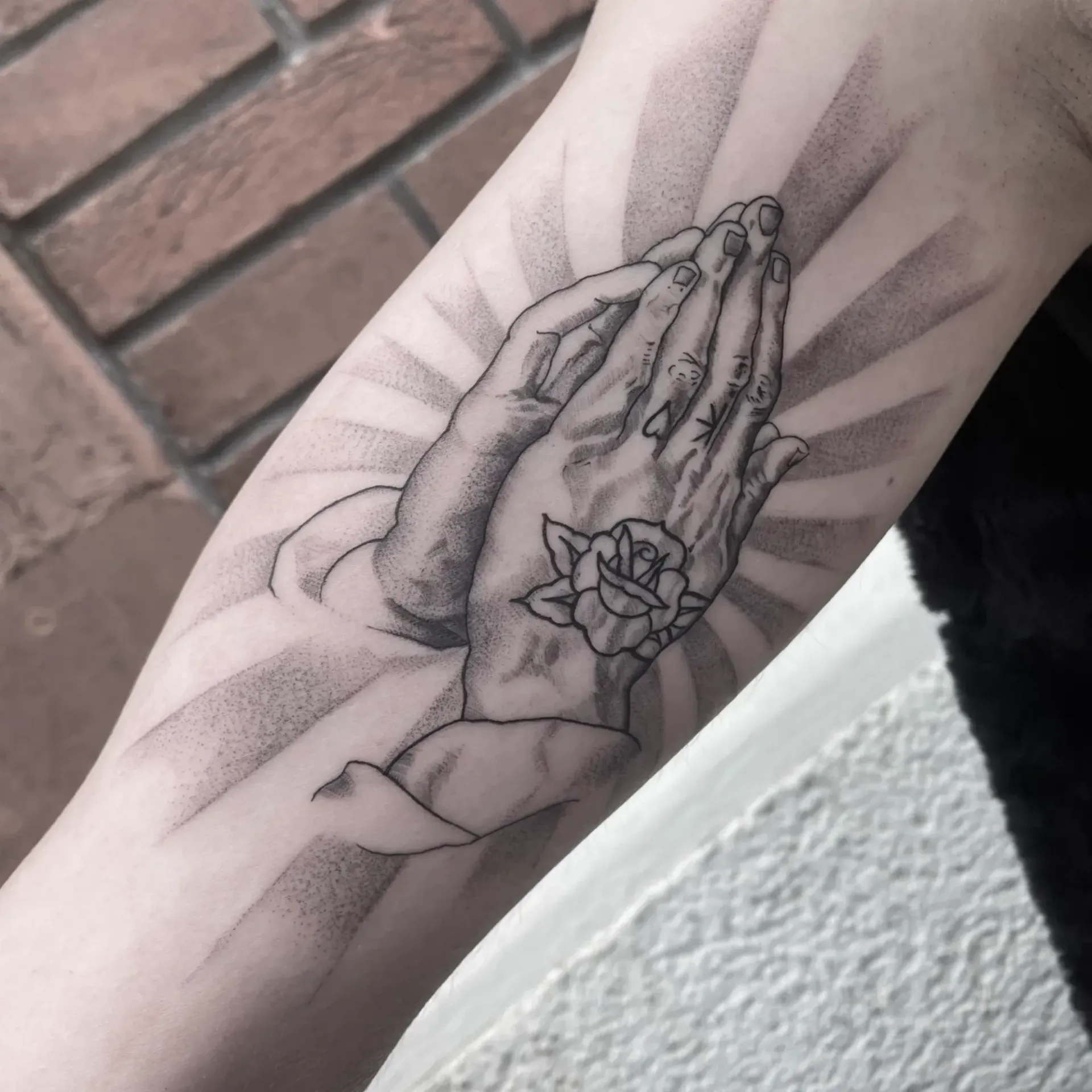 Un tatuaje en blanco y negro en un brazo que muestra unas manos rezando con una rosa en la muñeca, rodeadas de un sombreado que simula rayos de sol.
