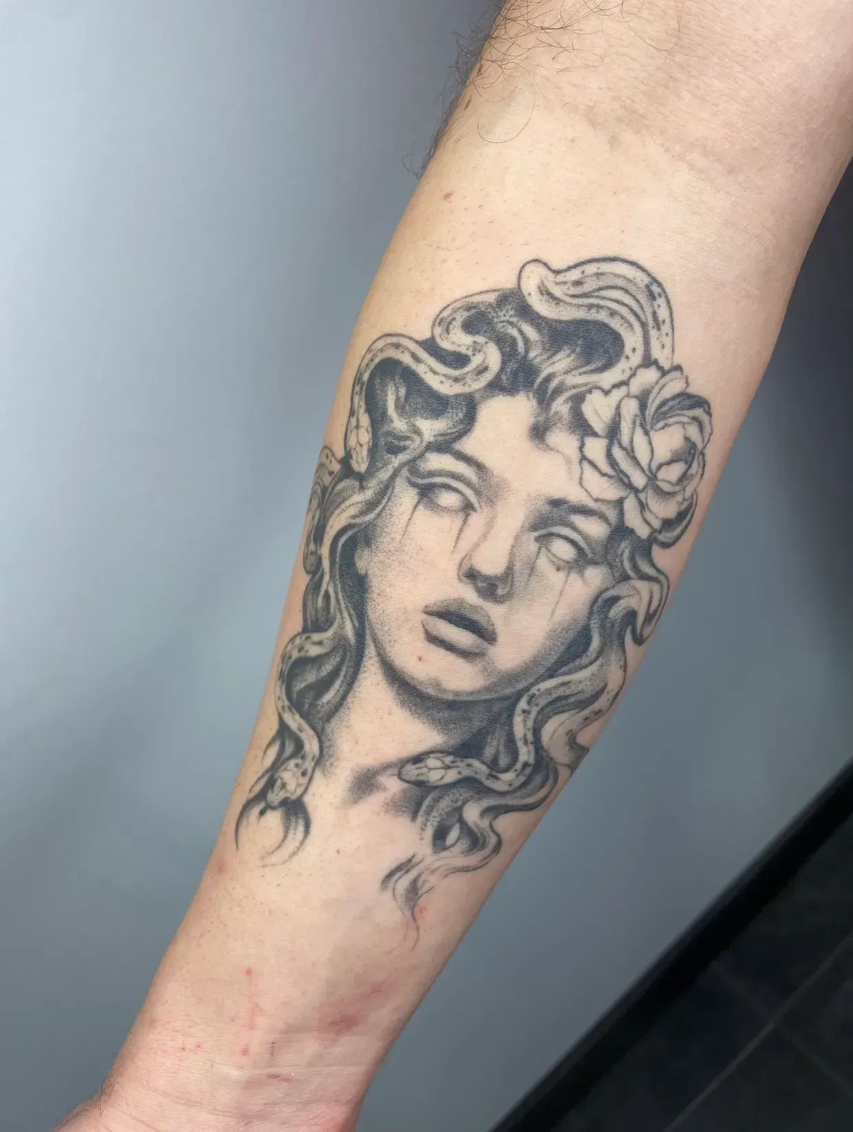 Tatuaje en blanco y negro en la parte interior del antebrazo que representa una figura estilizada de Medusa con serpientes en lugar de cabello y un detalle de rosa.