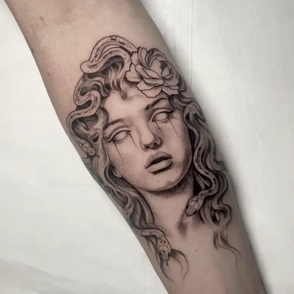 Un tatuaje detallado en blanco y negro de Medusa con cabello de serpiente, una rosa y ojos ciegos y llorosos en el antebrazo de una persona.