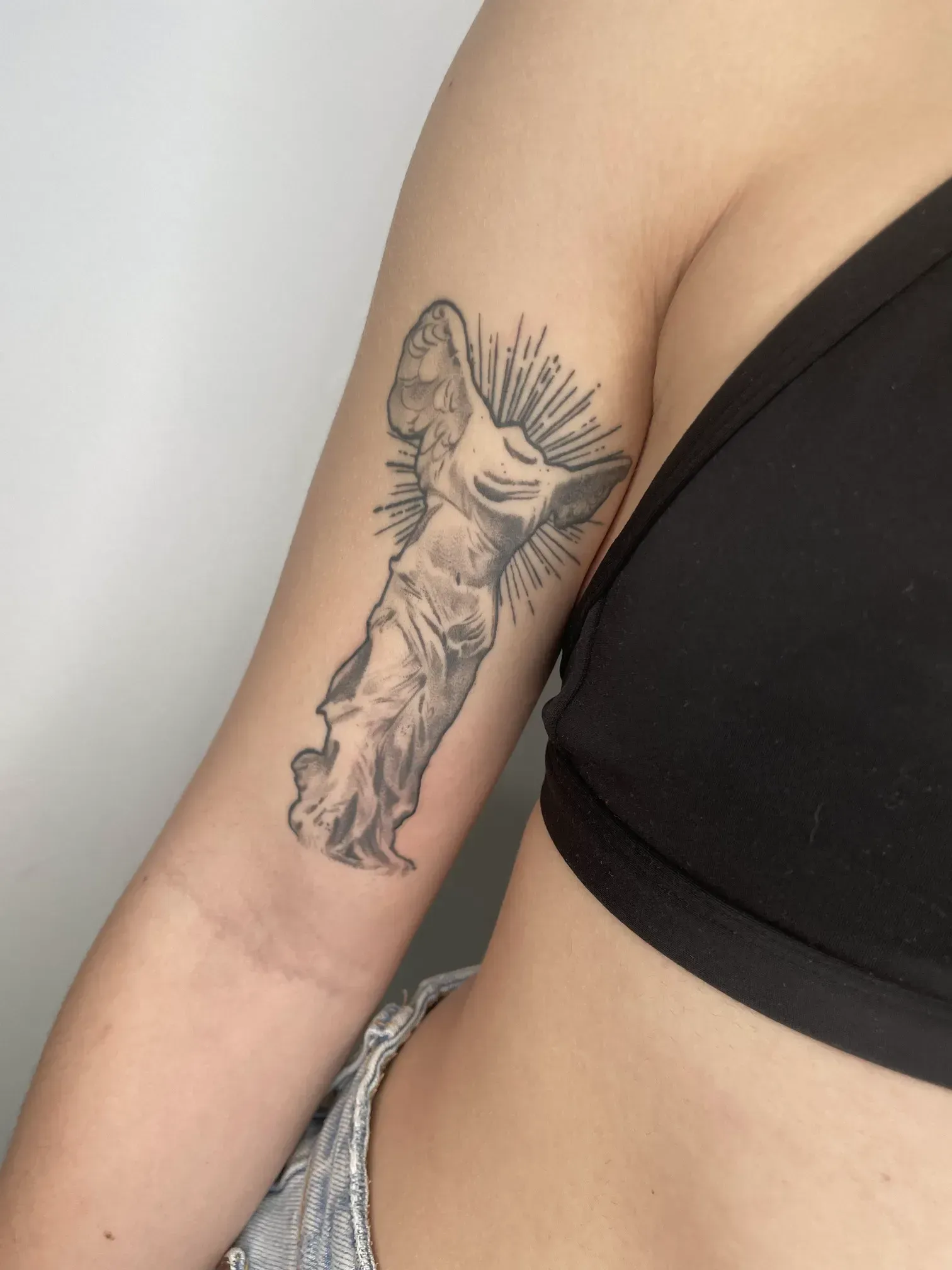 Un tatuaje en blanco y negro de la Victoria Alada de Samotracia en la parte superior del brazo, detallado con rayos de sol dibujados con líneas.