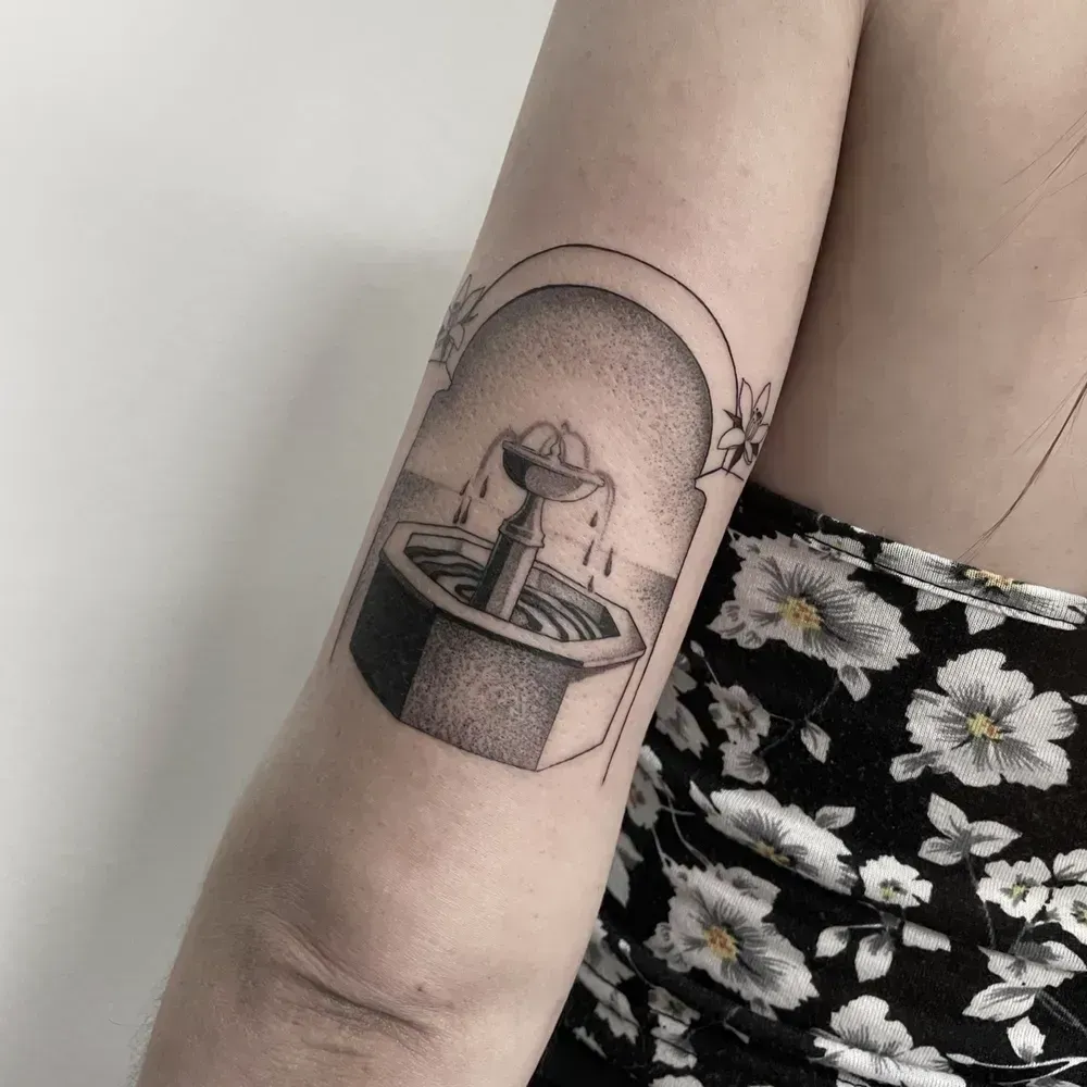 Tatuaje en la parte superior del brazo que representa una pequeña fuente en un marco arqueado, junto a un vestido negro estampado de flores.