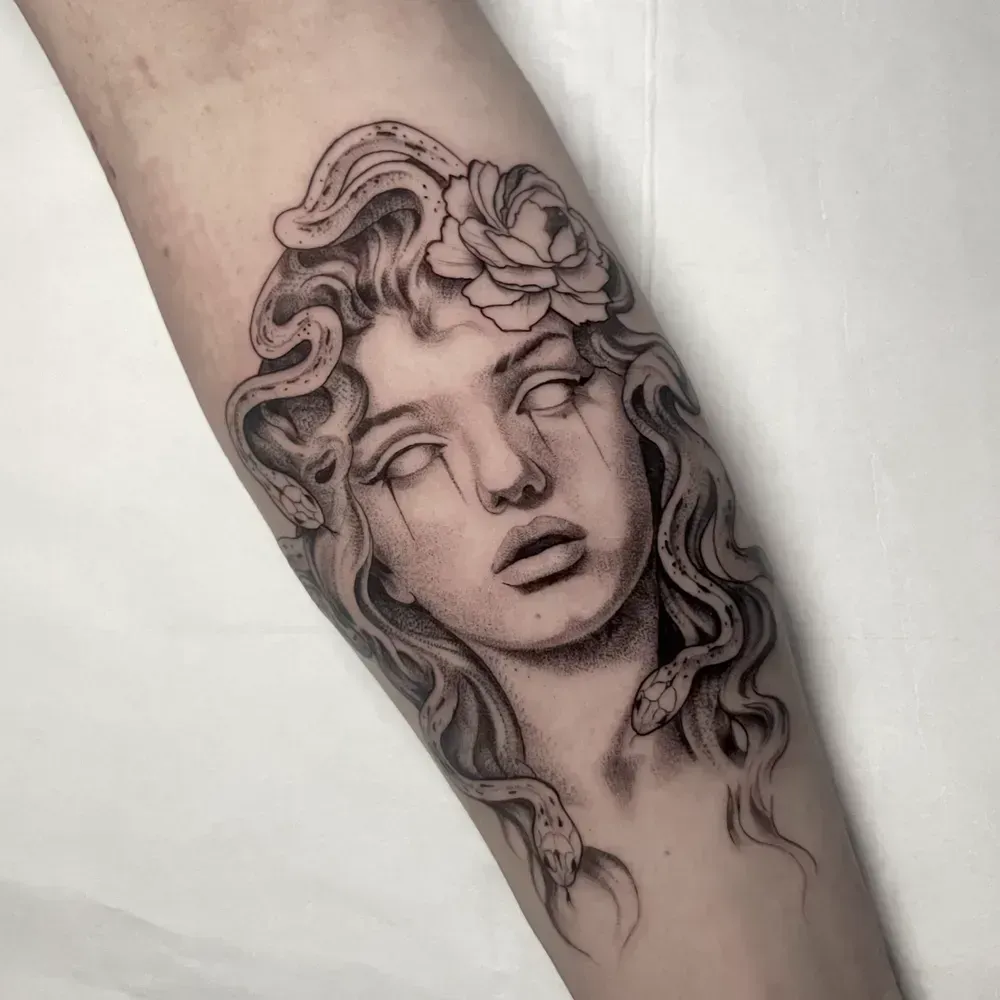 Tatuaje en blanco y negro de un rostro clásico con cabello suelto y una flor en el antebrazo.