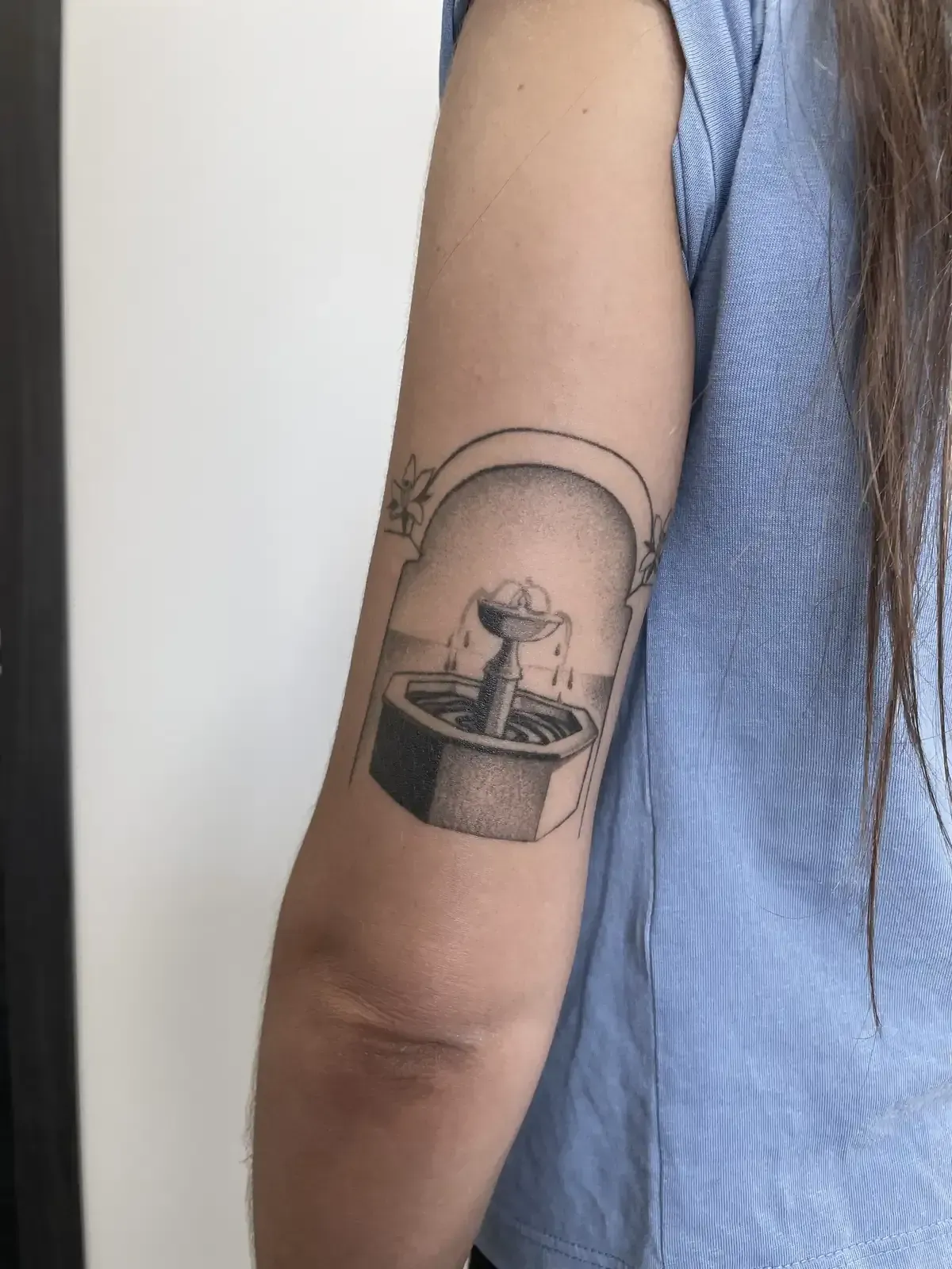 Un tatuaje de una pequeña fuente de piedra enmarcada en un arco en la parte posterior de un brazo.