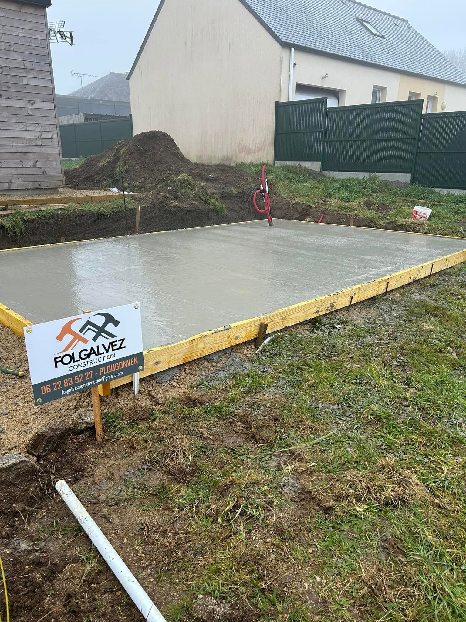Dalle de béton fraîche dans une cour avec un petit panneau de chantier et à proximité une clôture, une maison et du gazon.