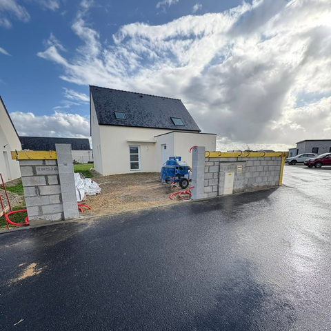 Maison neuve au toit en ardoise, piliers de portail en parpaing et allée détrempée sous un ciel nuageux.