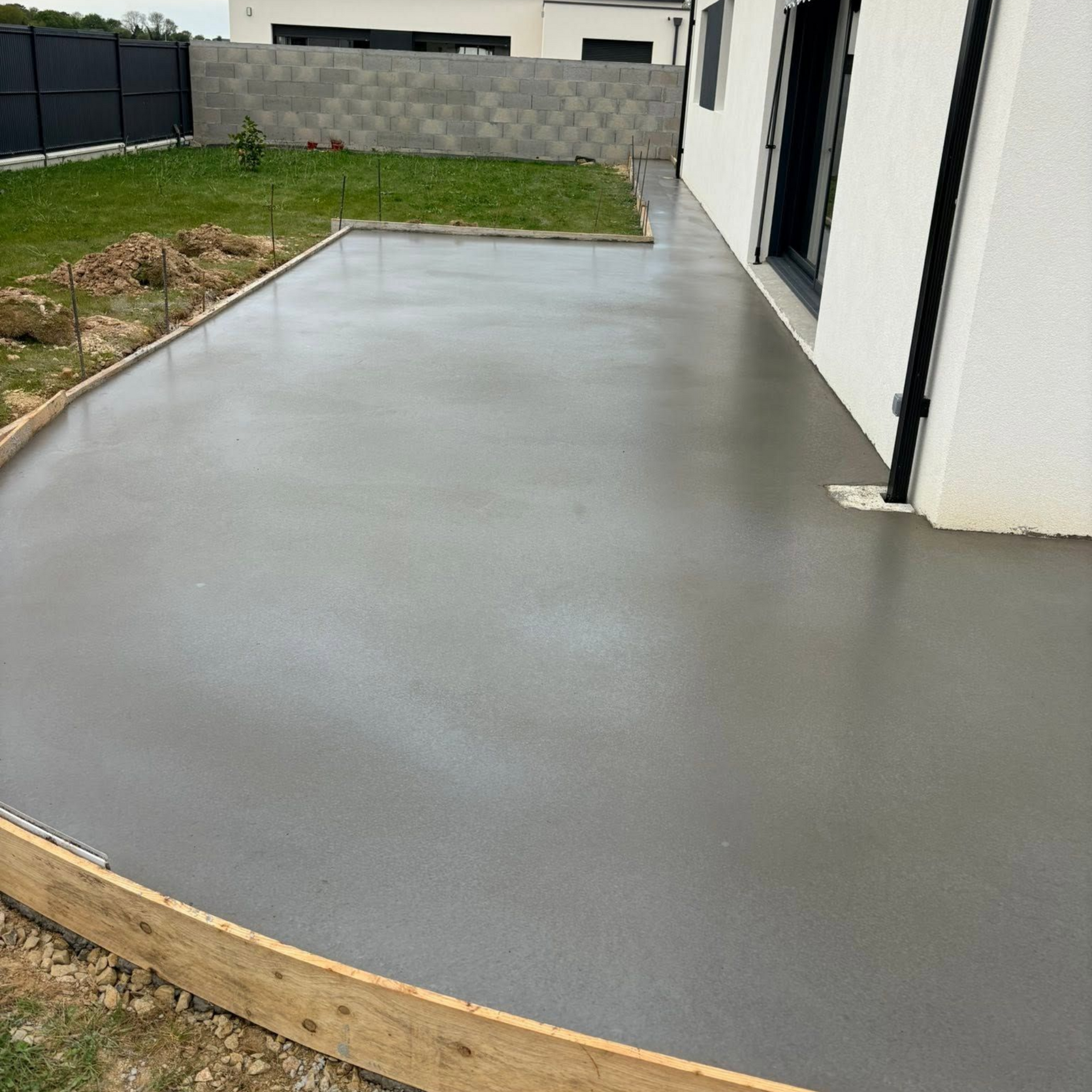 Terrasse en béton gris fraîchement coulée à côté d'une maison blanche avec un jardin gazonné et une clôture.
