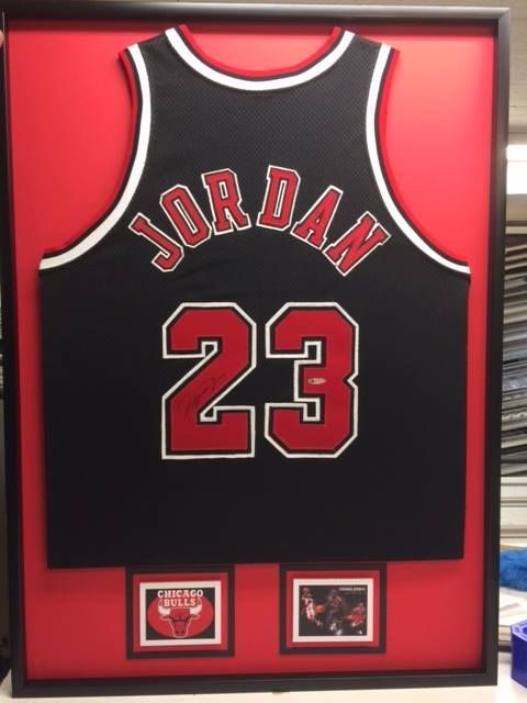 Kehyksiin pakattu Michael Jordan Chicago Bullsin pelipaita, musta punaisilla yksityiskohdilla, numero 23 ja kaksi pientä kehystettyä kuvaa.