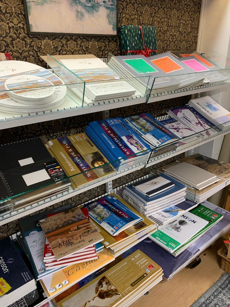 Hyllyt täynnä kirjoja ja paperitarvikkeita, kuten värikkäitä papereita, kortteja ja muistikirjoja, myymälämiljöössä.