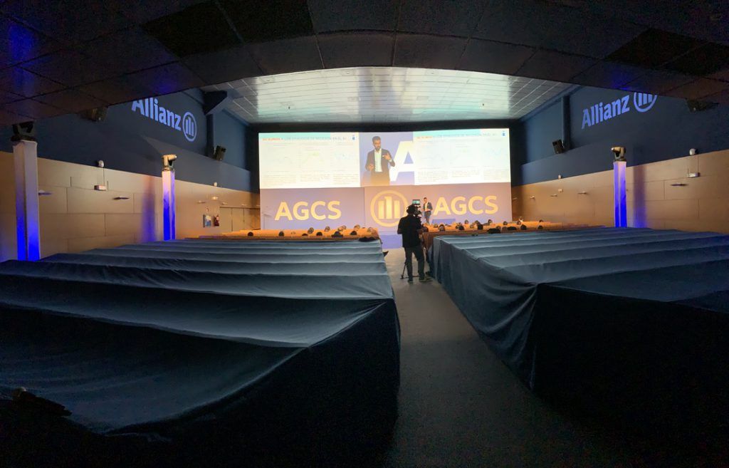 Auditorio vacío con filas de asientos tapizados de azul frente a un escenario y una pantalla iluminados