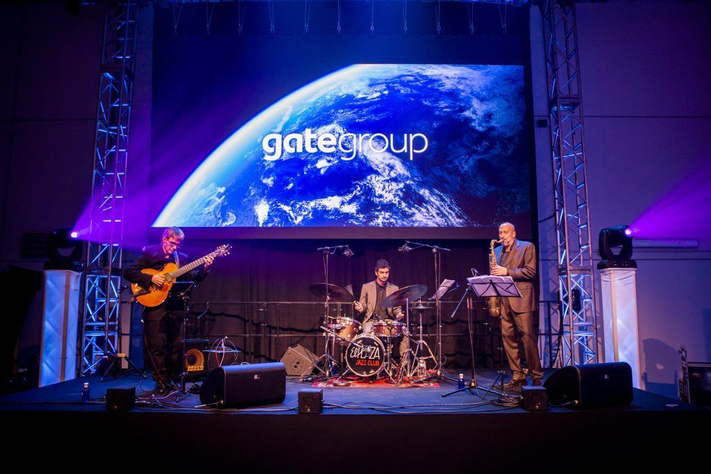Grupo musical en directo actuando en un escenario iluminado con luces moradas y con una gran pantalla de fondo