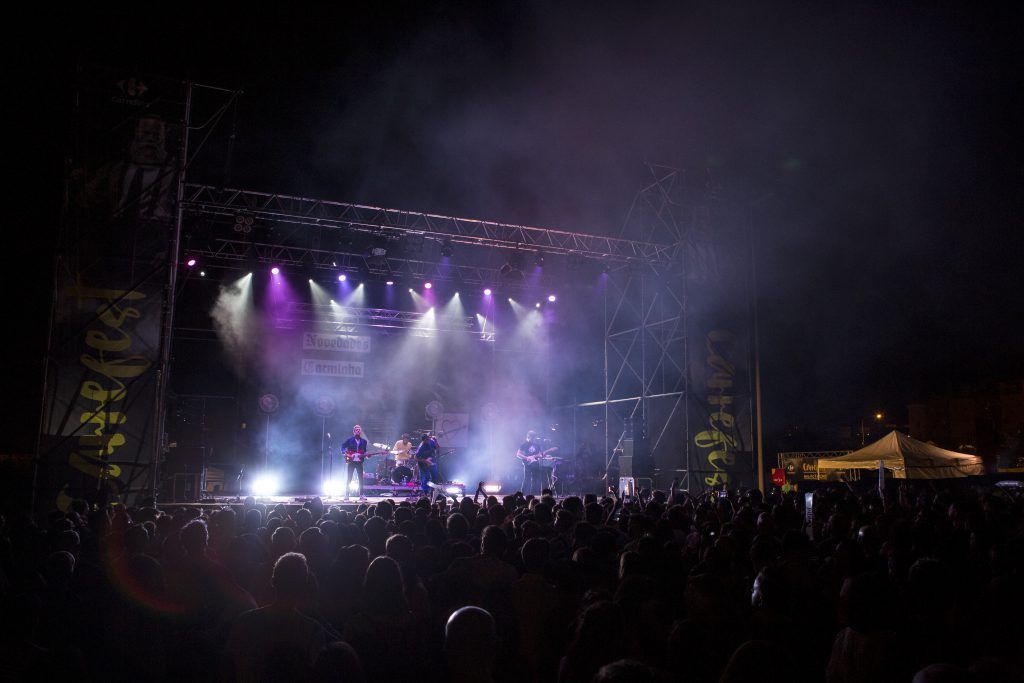 Escenario de concierto al aire libre iluminado por una neblina púrpura, con artistas y público frente a él.