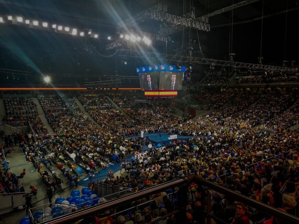Un recinto cubierto abarrotado con una gran multitud y un escenario iluminado para un mitin o evento político.