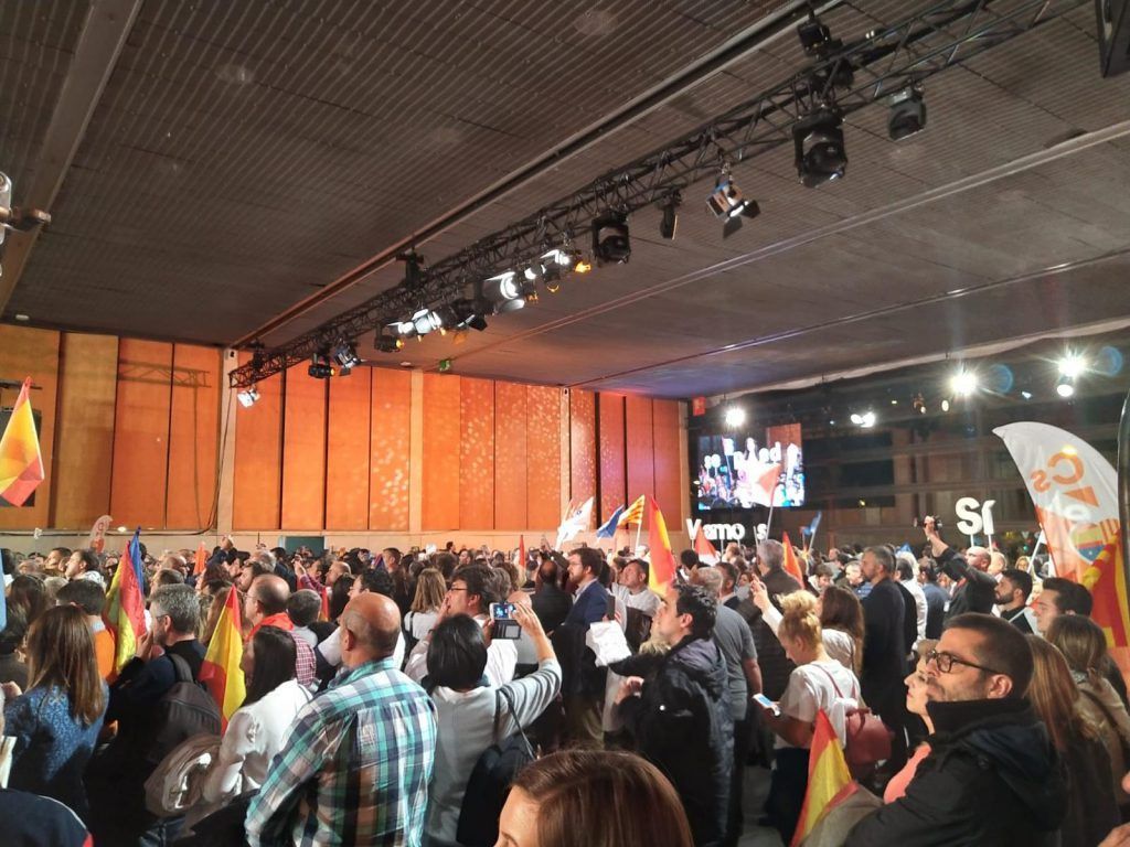 Multitud de personas en un evento en interiores, muchas levantando las manos bajo las luces del escenario